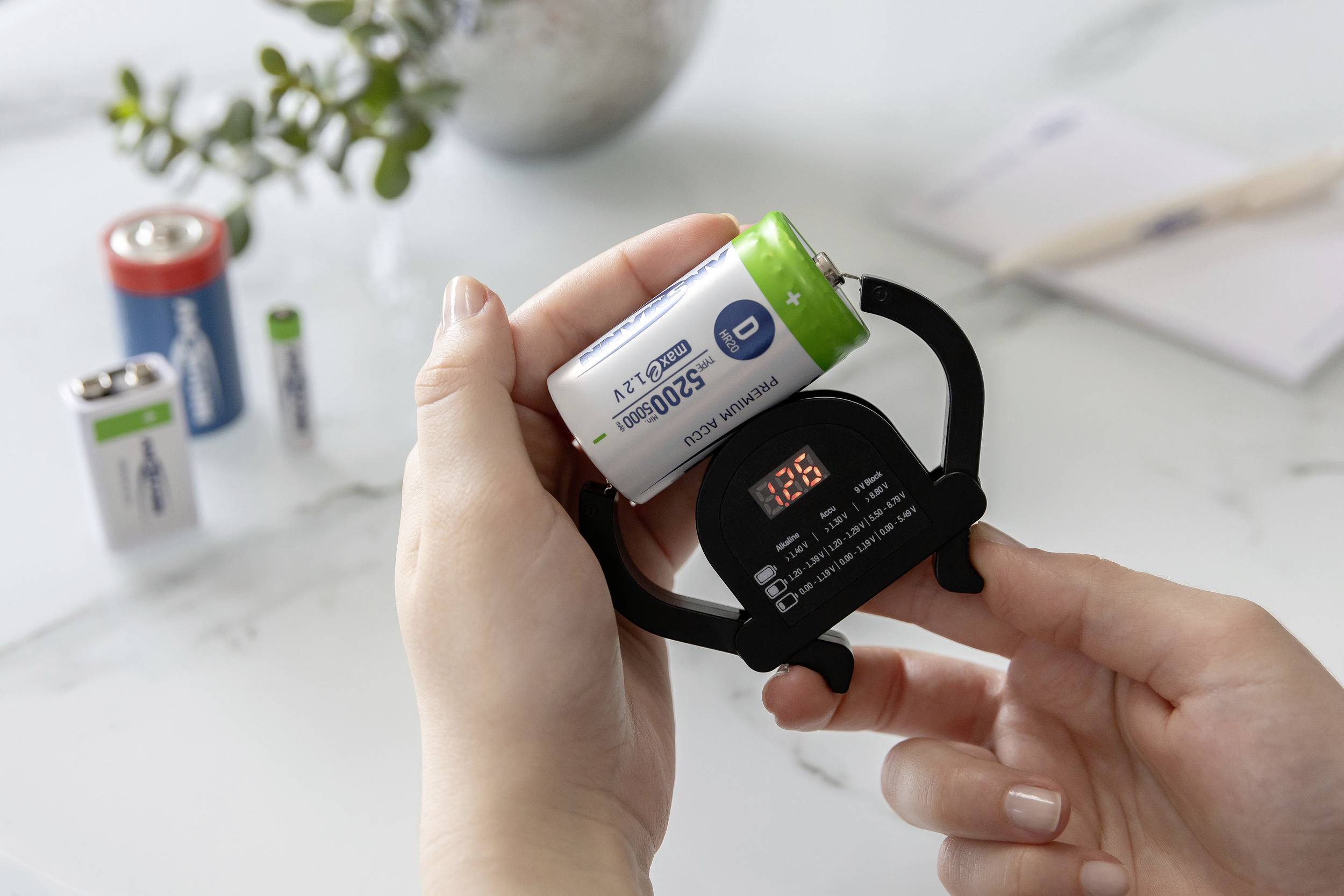 Person misst den Ladezustand einer Batterie mit einem Batterietester in der Hand; weitere Batterien im Hintergrund auf dem Tisch.
