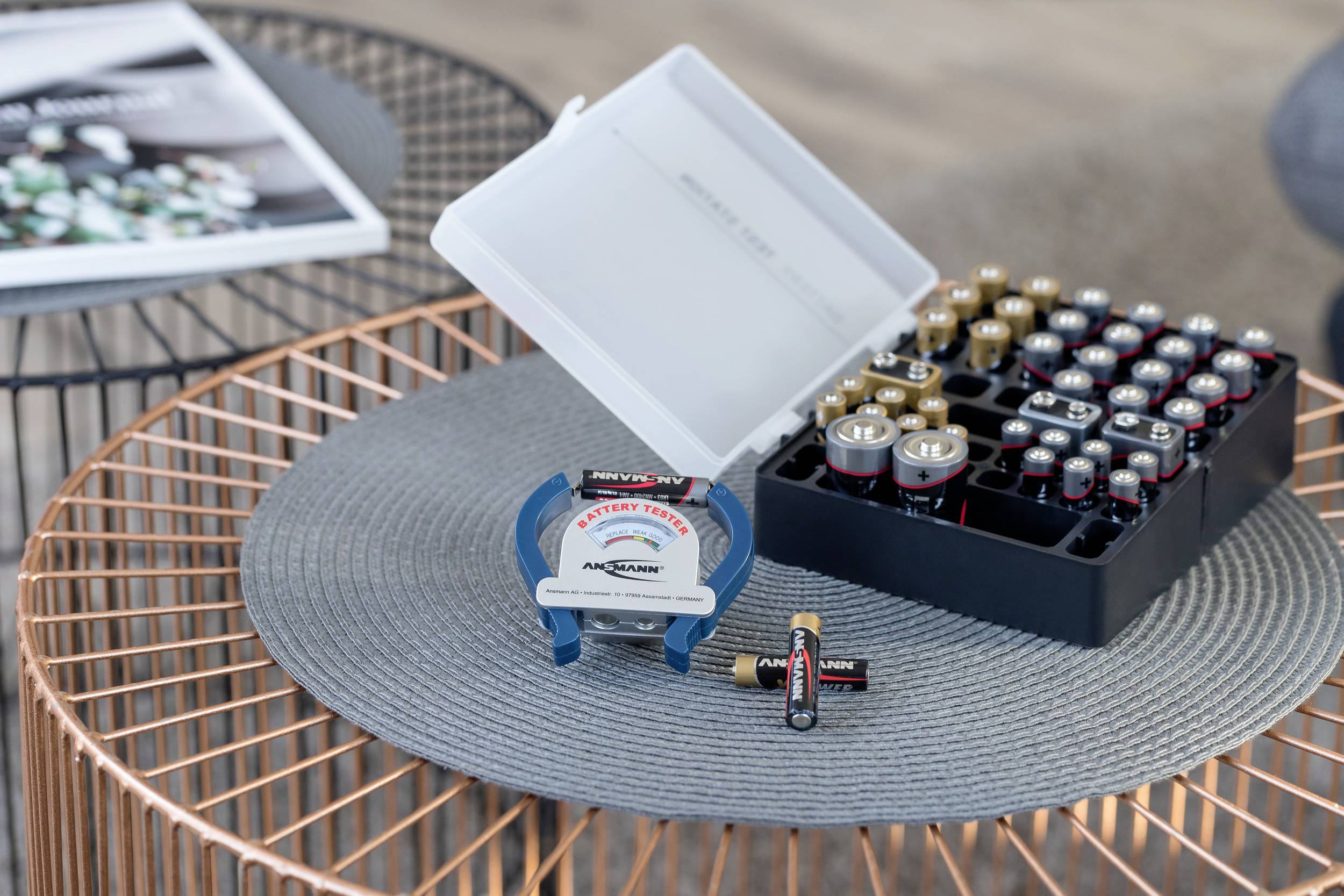 Ein Tisch mit einer runden Matte zeigt ein Batterieprüfgerät, Batterien in einem Kasten und einige lose Batterien in der Nähe.