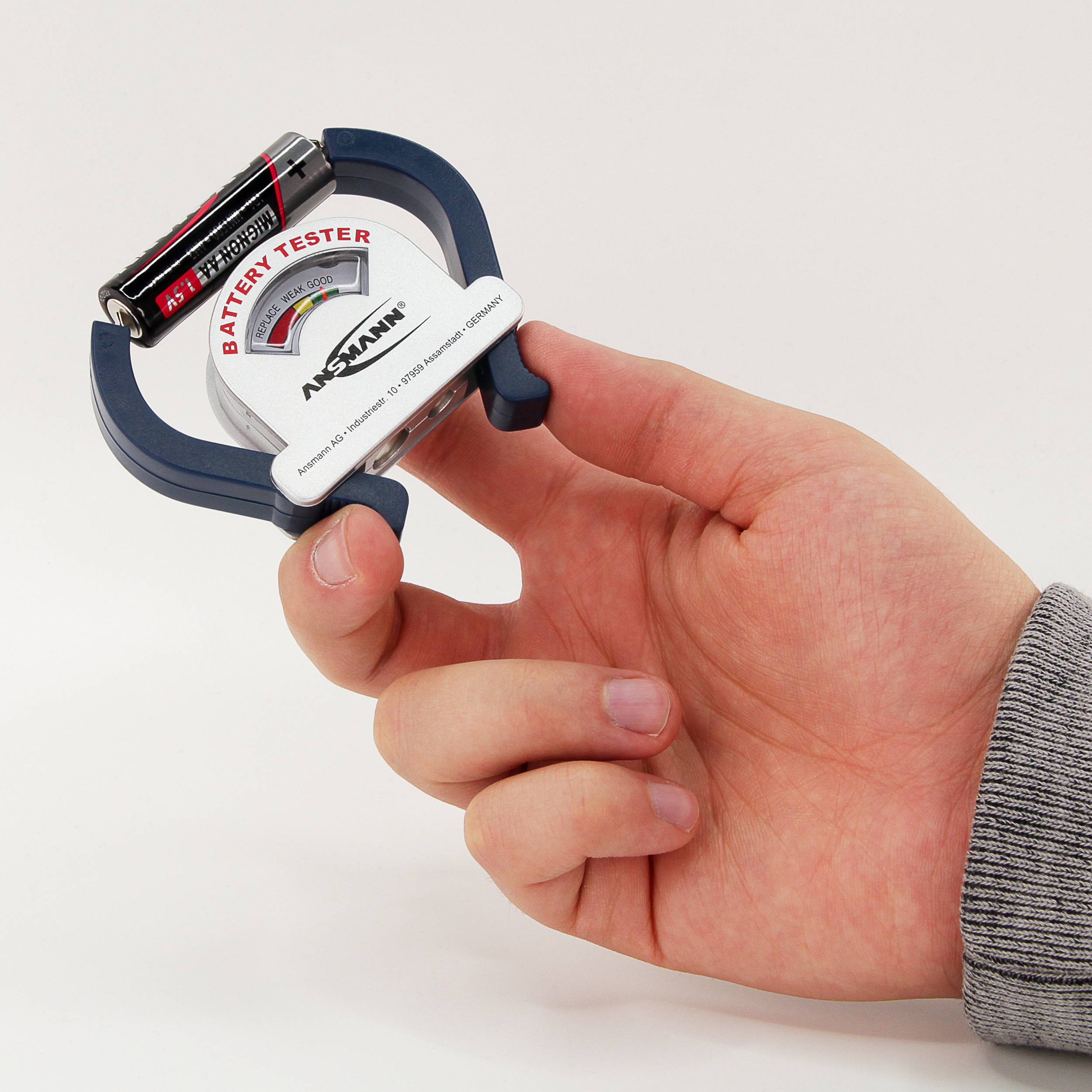 Eine Hand hält einen Batterietester mit einer eingelegten AA-Batterie. Der Tester zeigt den Batteriestand mit einem Farbdiagramm an.