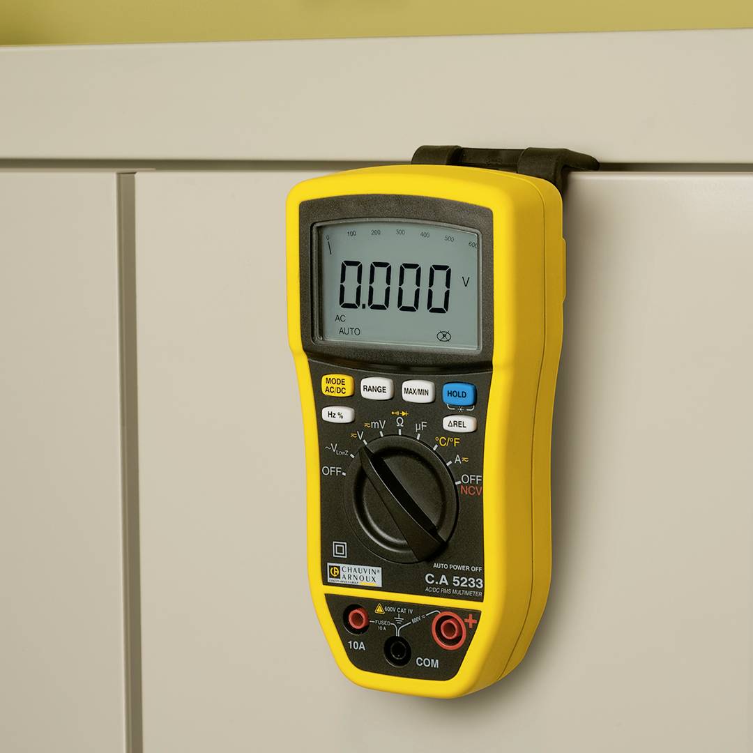 Ein gelbes digitales Multimeter hängt an einem weißen Schrank. Display zeigt '0.000 V'. Verschiedene Messfunktionen sind wählbar.