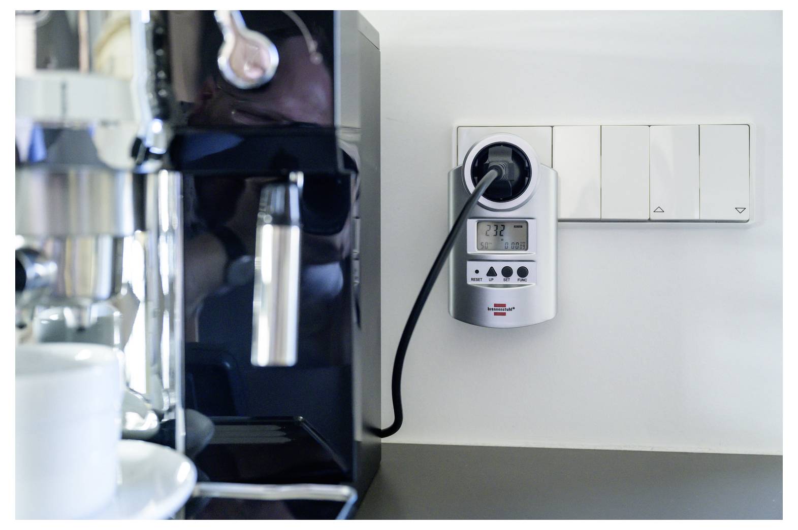 Elektrokaffeemaschine mit einem Stecker-Strommessgerät, das den Energieverbrauch anzeigt, neben Wandschaltern in einer Küchen- oder Café-Umgebung.