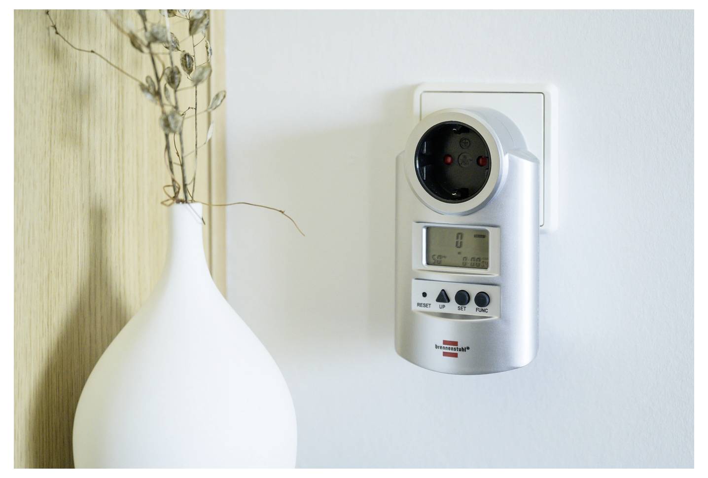 An der Wand montiertes digitales Timer-/Steuergerät neben einer weißen Vase mit getrockneten Zweigen auf einem Regal.