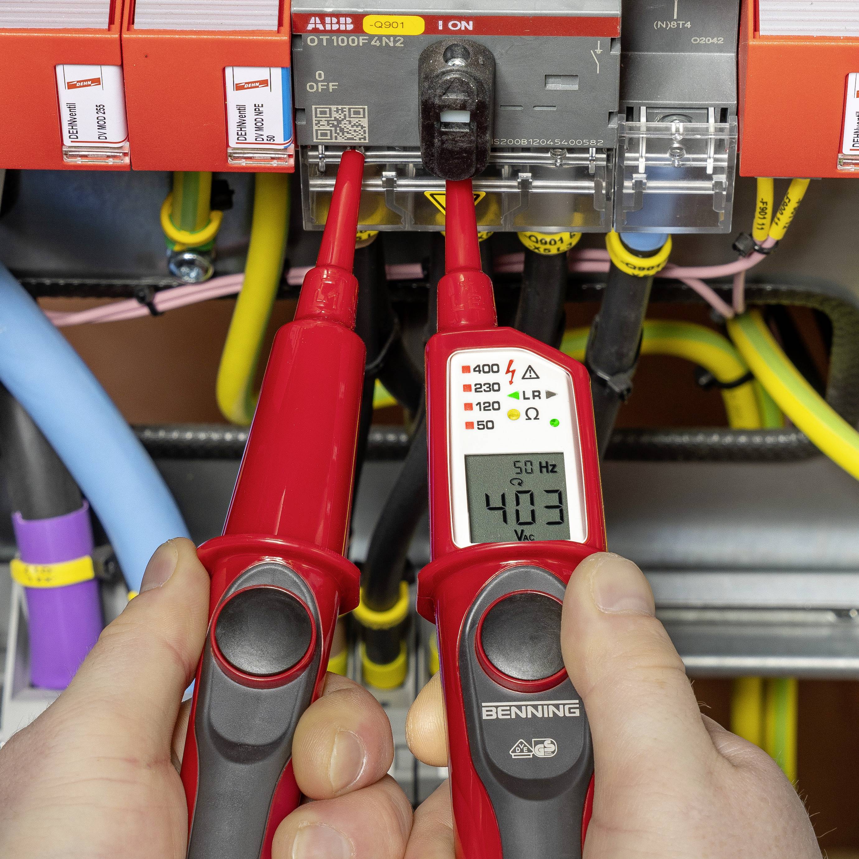 Zwei Hände halten rote Messgeräte, die an einen Schaltkasten angeschlossen sind, und zeigen eine Messung von 403 Hz und 230 V.