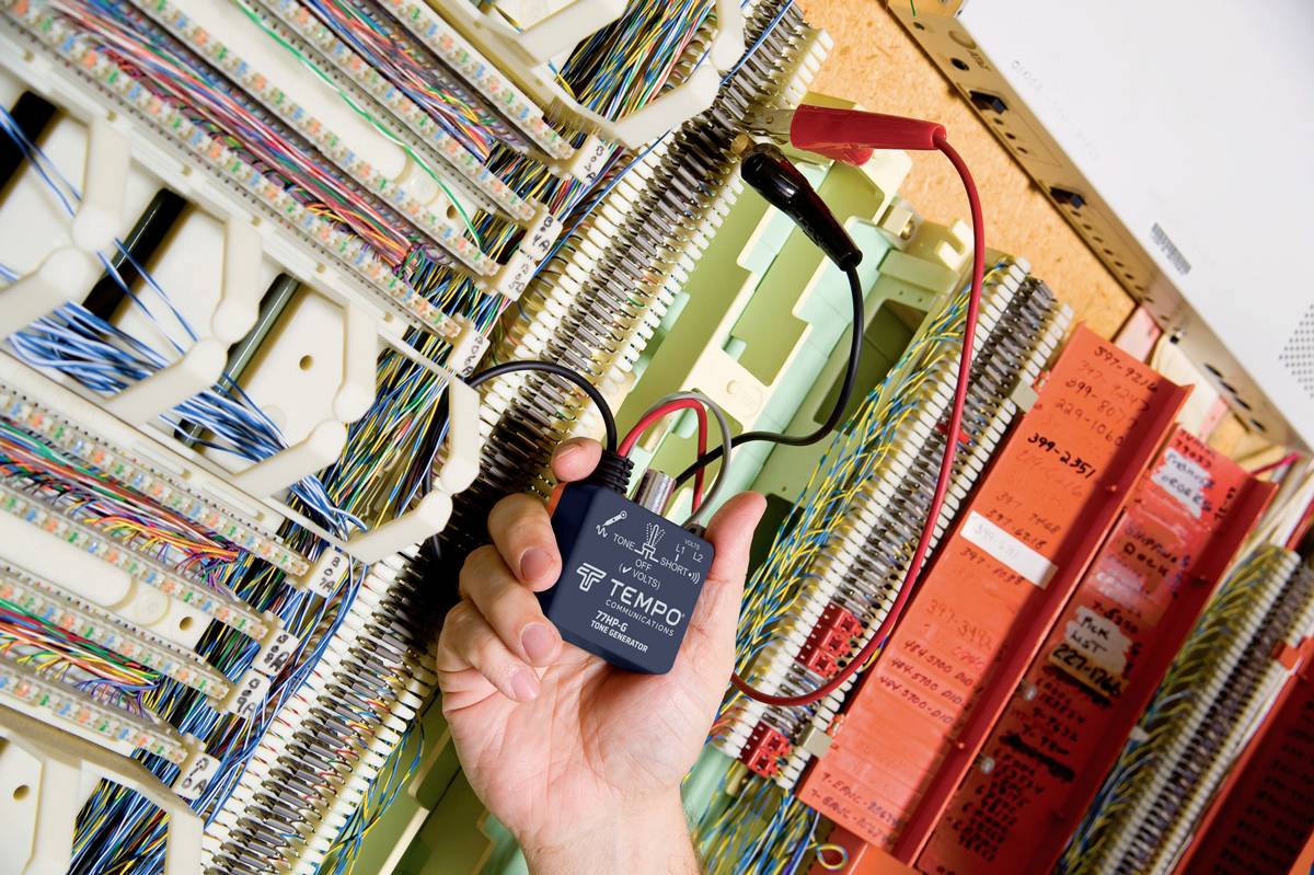 Eine Person hält ein schwarzes Gerät mit Kabeln in der Hand, umgeben von zahlreichen Telefonstrippen in einem Schaltkasten.