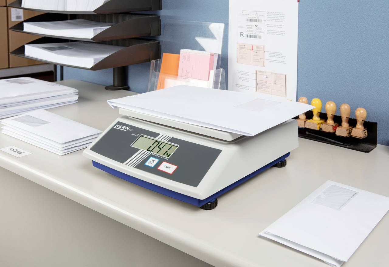 Eine digitale Waage zeigt '0.44 kg' an und wiegt einen Stapel Briefe. Im Hintergrund sind Briefmarken und weitere Umschläge sichtbar.