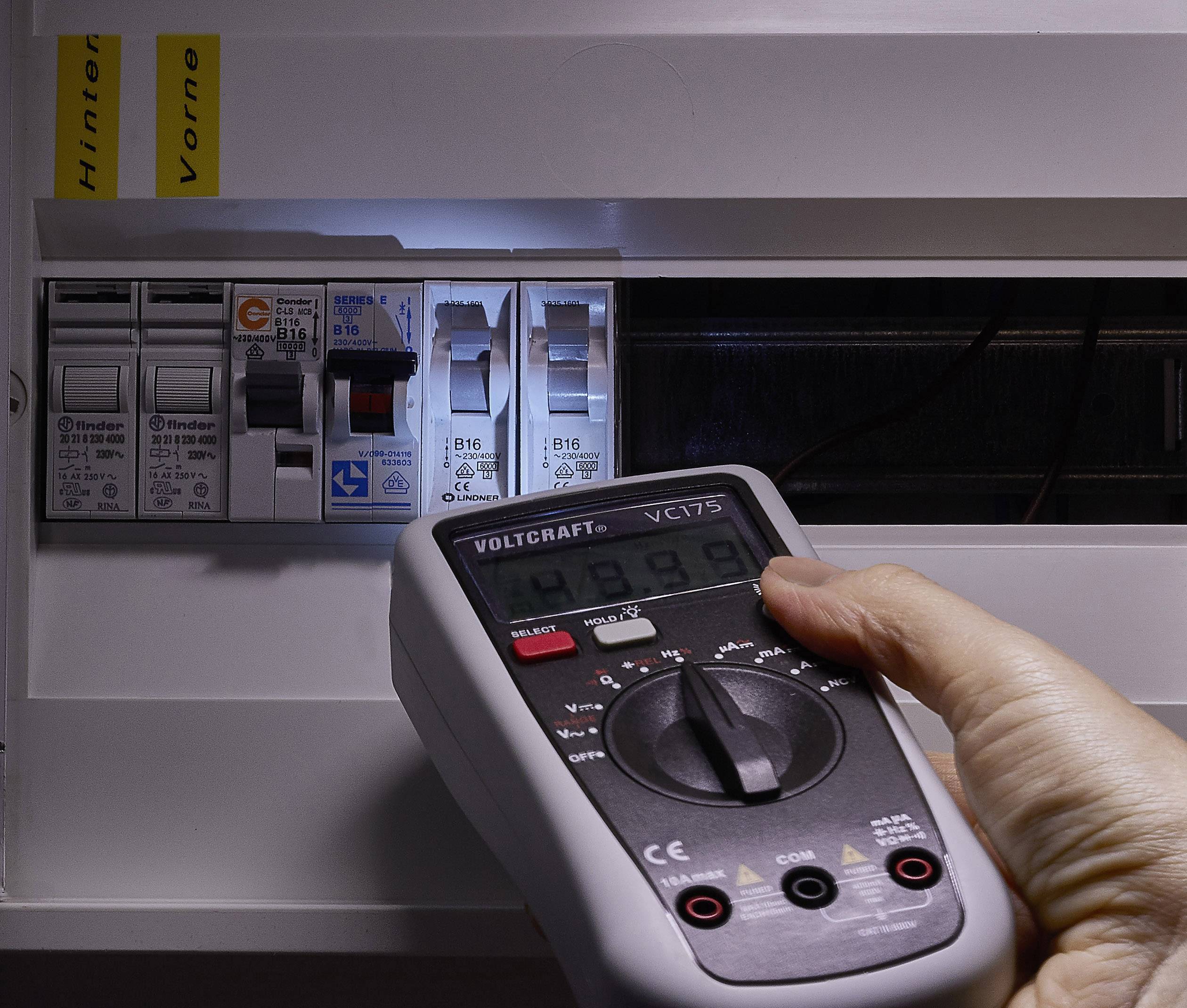 Hand hält ein Multimeter vor einem geöffneten Sicherungskasten, prüft die Spannungswerte. Der Fokus liegt auf Präzision und Sicherheit.