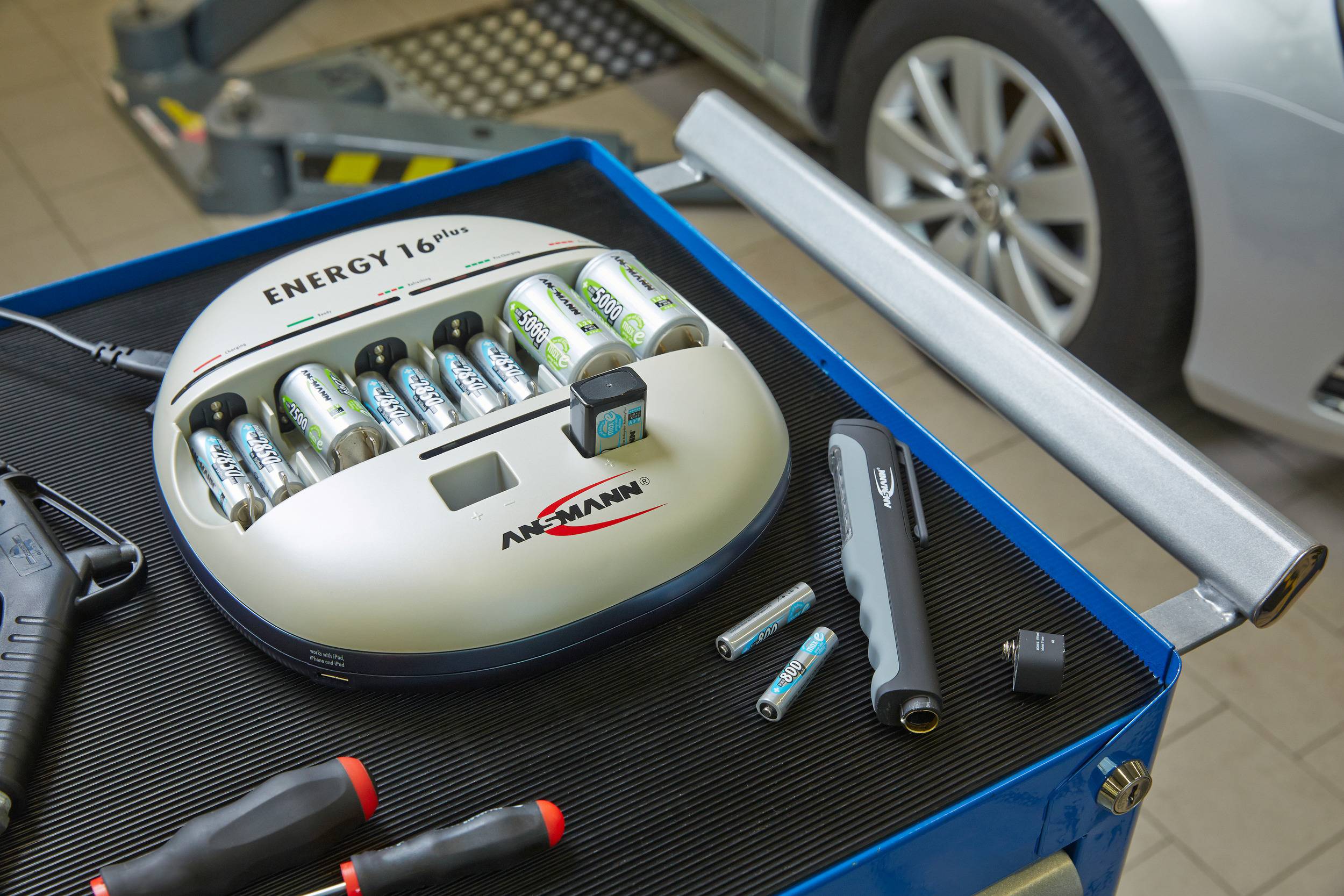 Batterieladegerät auf Werkbank mit geladenen Batterien, daneben Werkzeuge. Auto im Hintergrund. Werkstattumgebung.