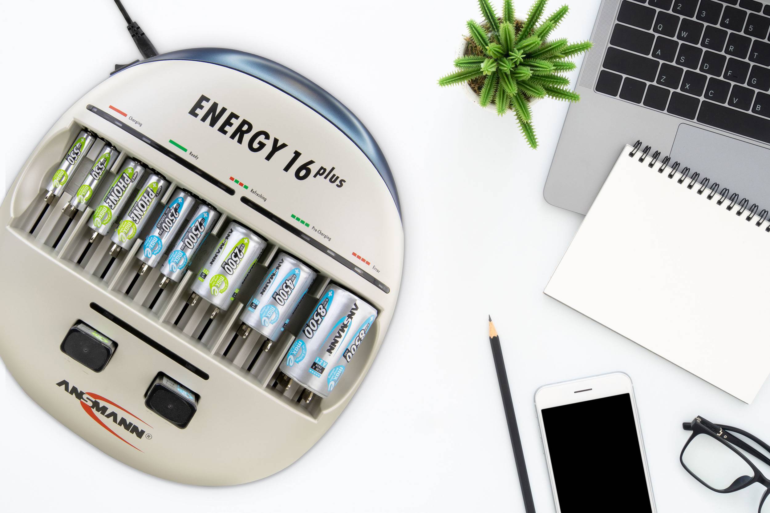 Akkuladegerät mit sechs eingelegten AA-Batterien neben einem Laptop, einem Notizbuch, einem Smartphone und einer kleinen Pflanze.