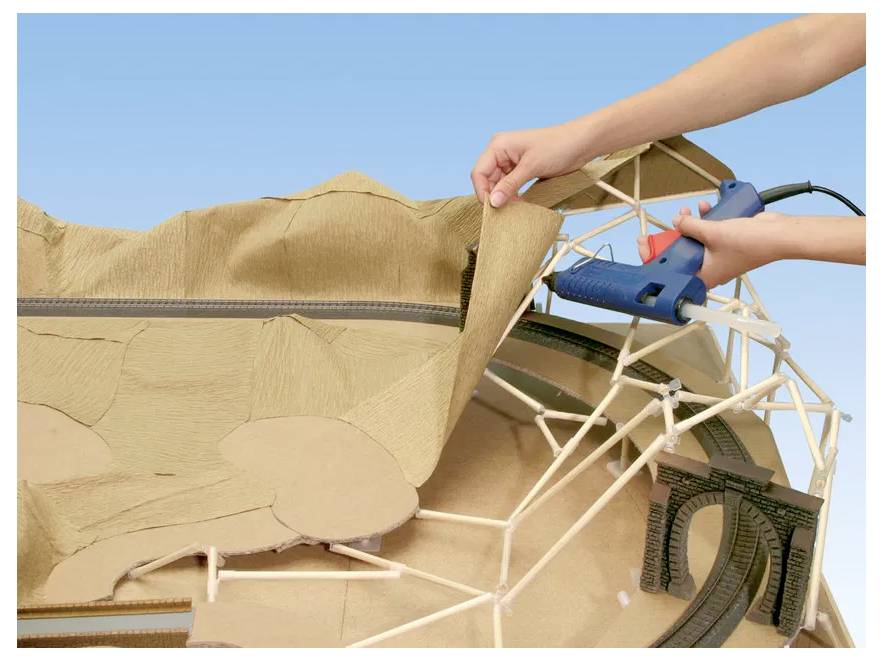 A person uses a glue gun to attach brown textured paper to a white 3D model structure, possibly crafting a landscape scene.