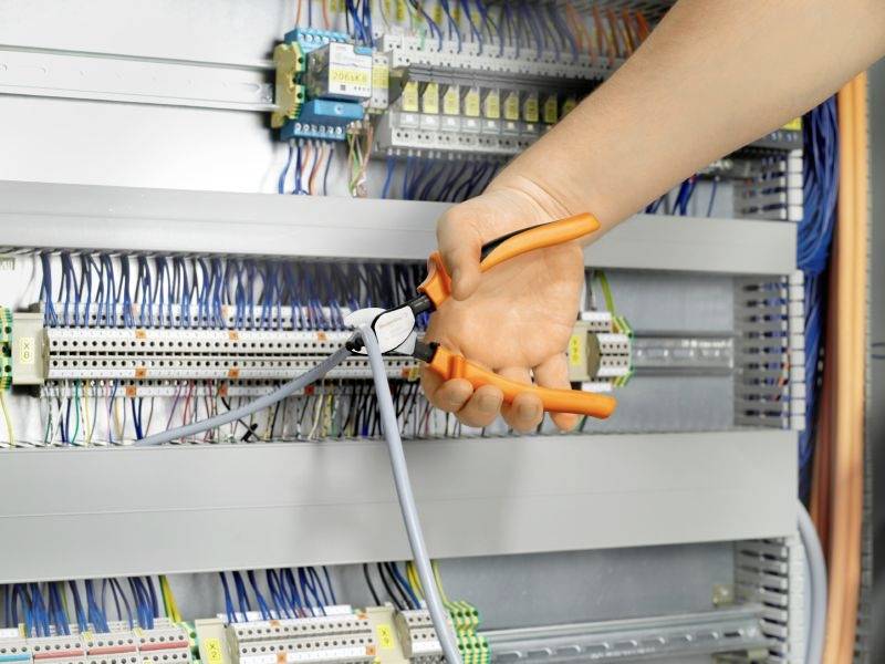 Eine Hand hält orangefarbene Zangen, die ein graues Kabel in einem Schaltschrank durchtrennen. Verlegte Drähte und Bauteile sind sichtbar.