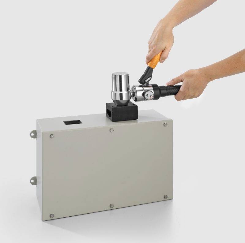 Eine Person, die ein Werkzeug verwendet, um eine metallische Komponente an einer großen rechteckigen grauen Box vor einem einfarbigen Hintergrund einzustellen.