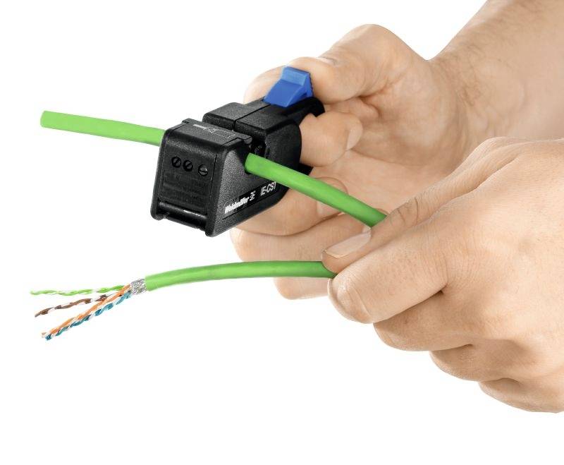 Hände halten ein Entmantelungswerkzeug, das ein grünes Kabel entmantelt. Die inneren Drähte des Kabels sind sichtbar.