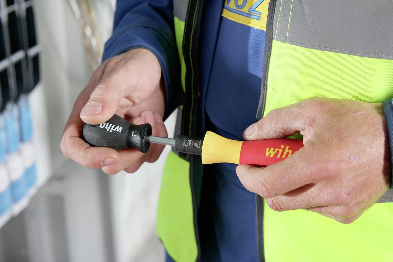 Eine Person hält zwei Schraubendreher mit schwarzen und roten Griffen in den Händen. Hintergrund unscharf mit technischer Ausrüstung.