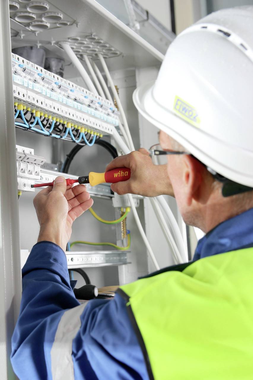 Ein Elektriker mit Schutzhelm und Warnweste arbeitet an einem Schaltschrank und nutzt einen Schraubendreher, um Verbindungen vorzunehmen.