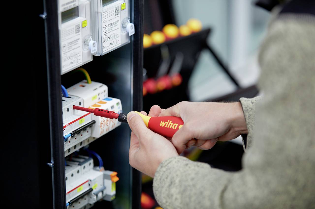 Eine Person benutzt einen Schraubendreher, um an einer elektrischen Schalttafel zu arbeiten. Im Hintergrund sind Werkzeuge zu sehen.