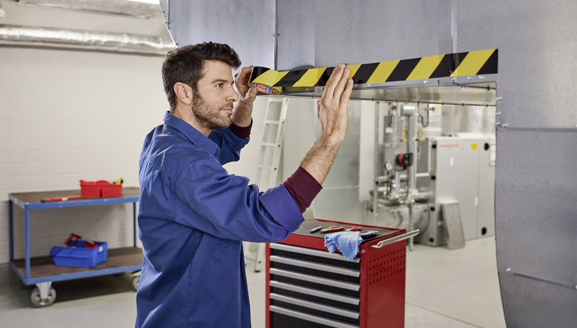 Ein Mann in Arbeitskleidung bringt ein Warnband an einer niedrigen Metallkante in einem industriellen Raum an. Im Hintergrund sind Werkzeuge zu sehen.