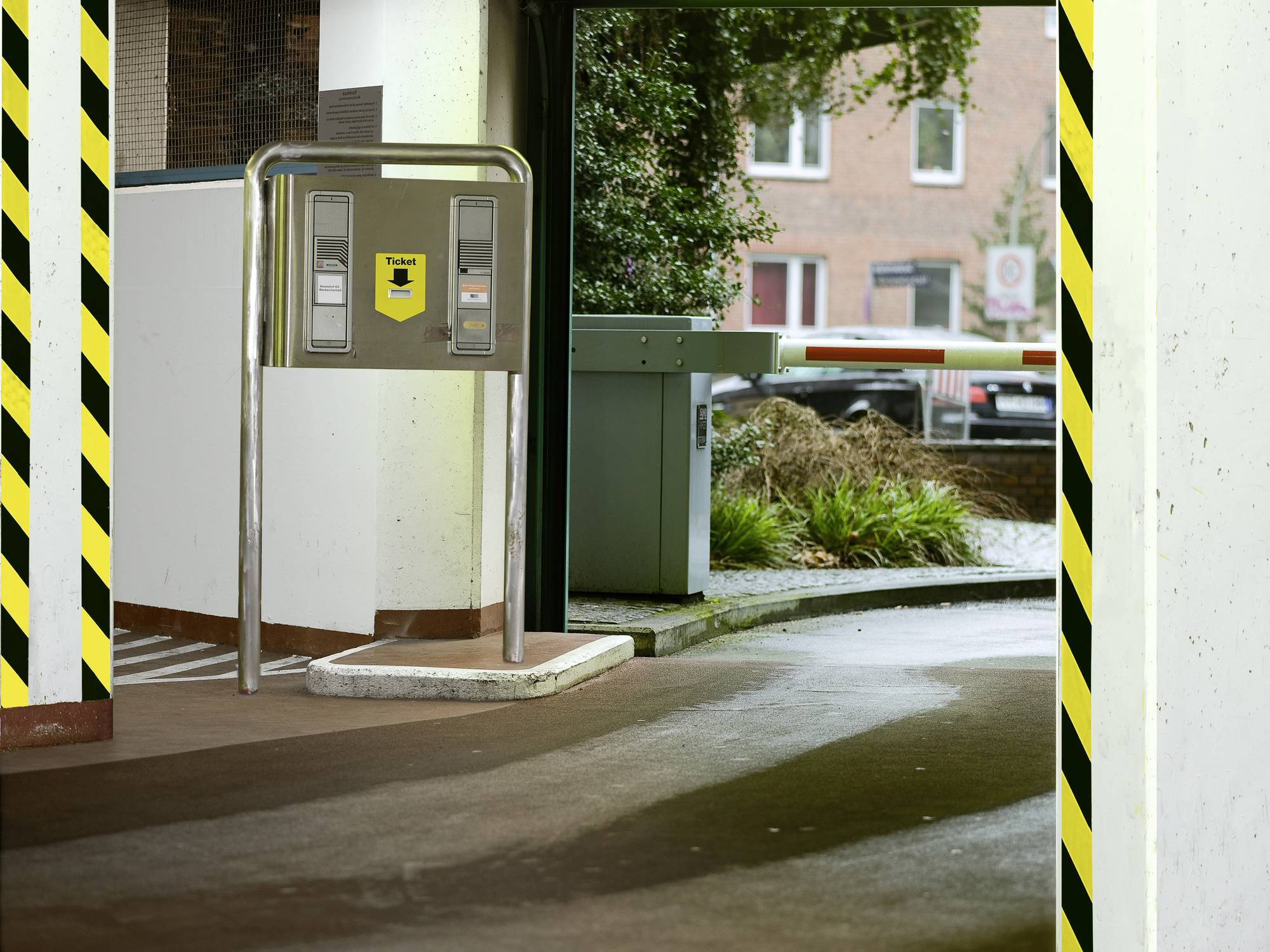 Parkhaus-Einfahrt mit Schranke und Ticketautomat. Im Hintergrund Bäume und Ziegelgebäude. Verkehrszeichen sichtbar.