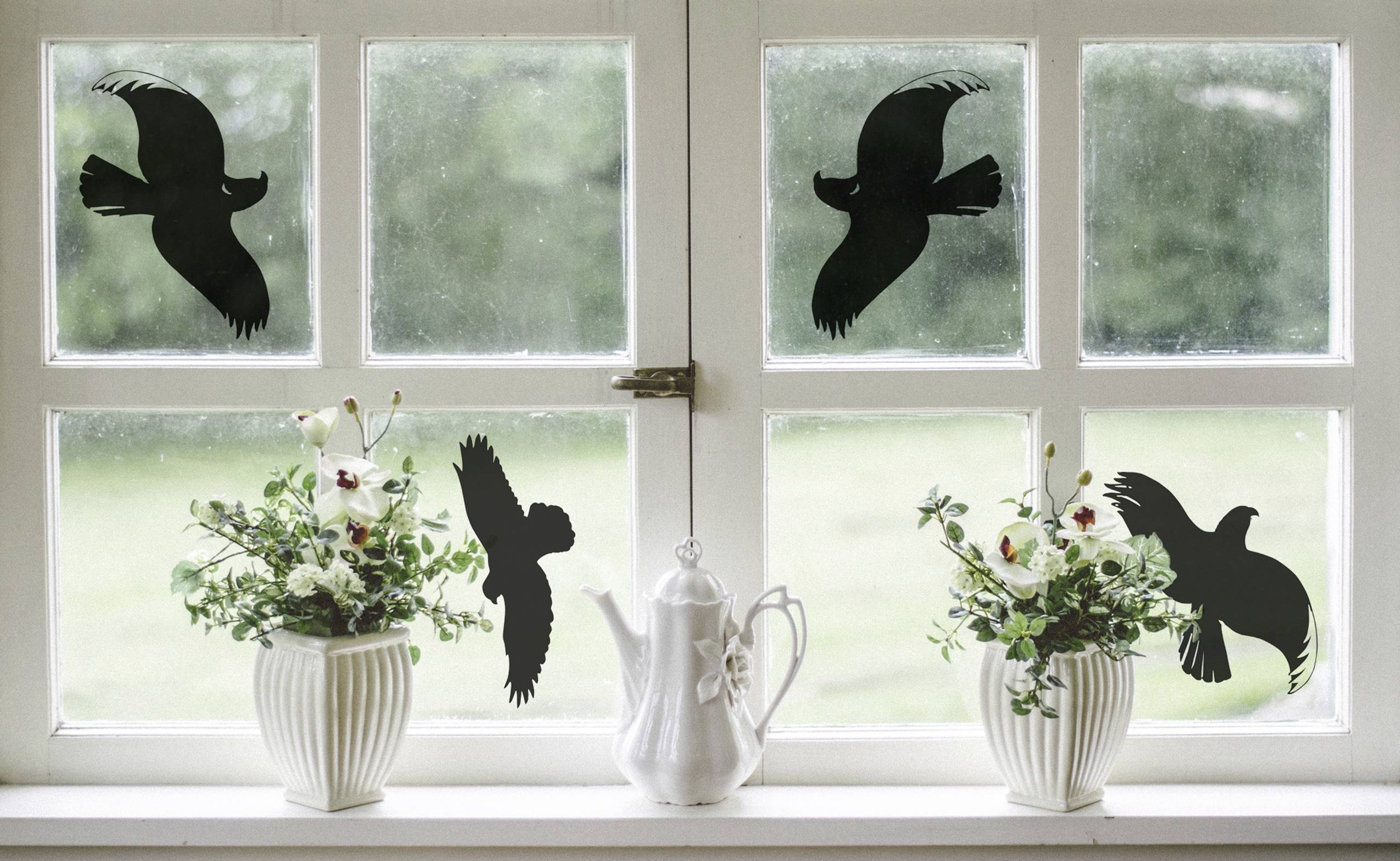 Ein Fenster mit Vogelaufklebern auf den Glasscheiben, flankiert von zwei weißen Vasen mit Blumen und einer weißen Teekanne in der Mitte.