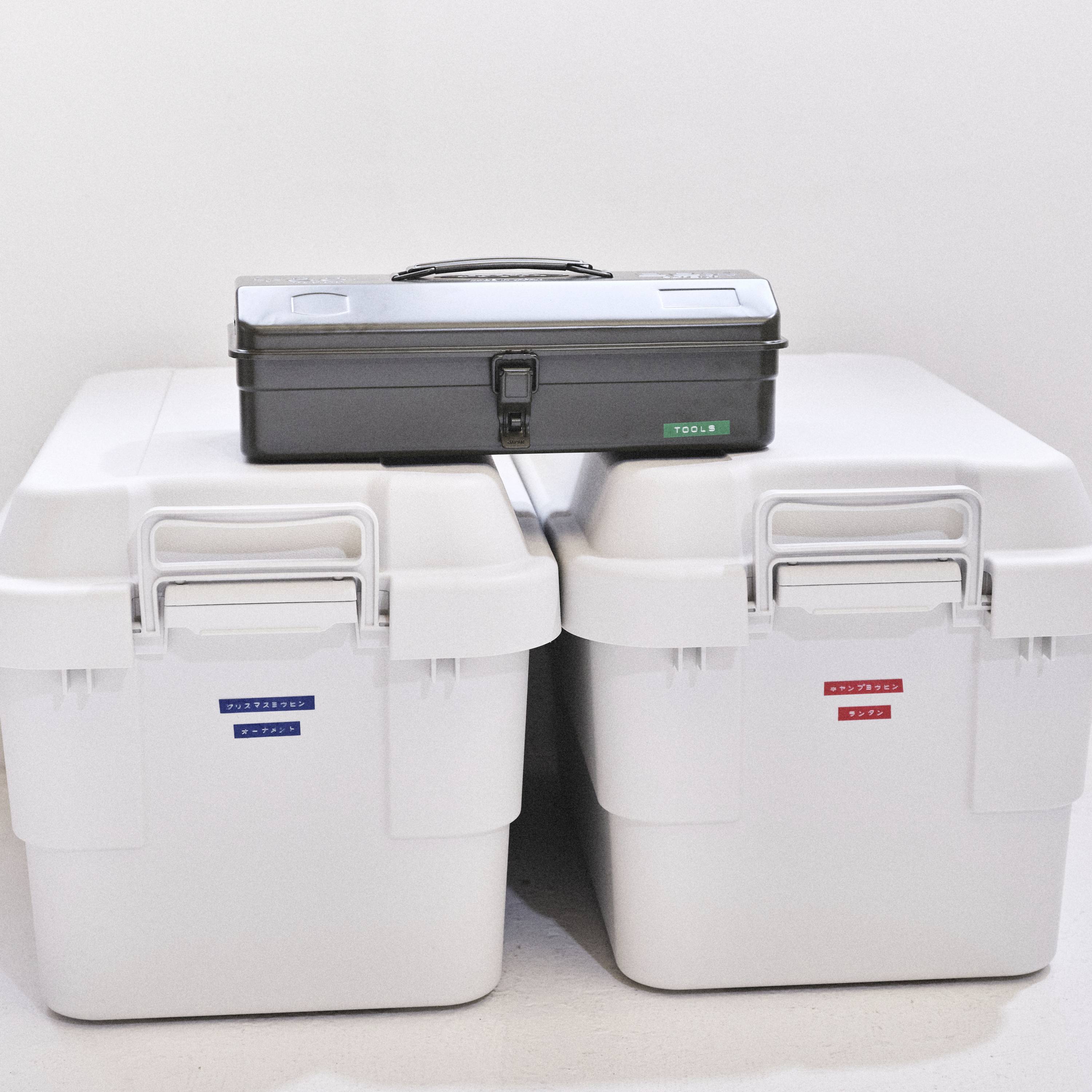 Ein schwarzer Werkzeugkasten steht auf zwei weißen Kisten mit Etiketten in Blau und Rot, die vor einer weißen Wand platziert sind.