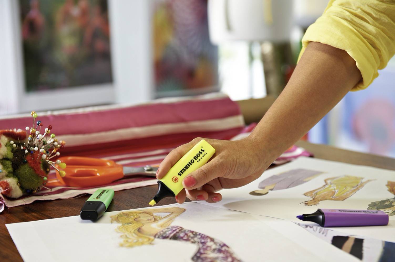 Eine Person markiert Modedesigns mit Textmarker in verschiedenen Farben. Neben einem Nadelkissen und Stoff auf dem Tisch.