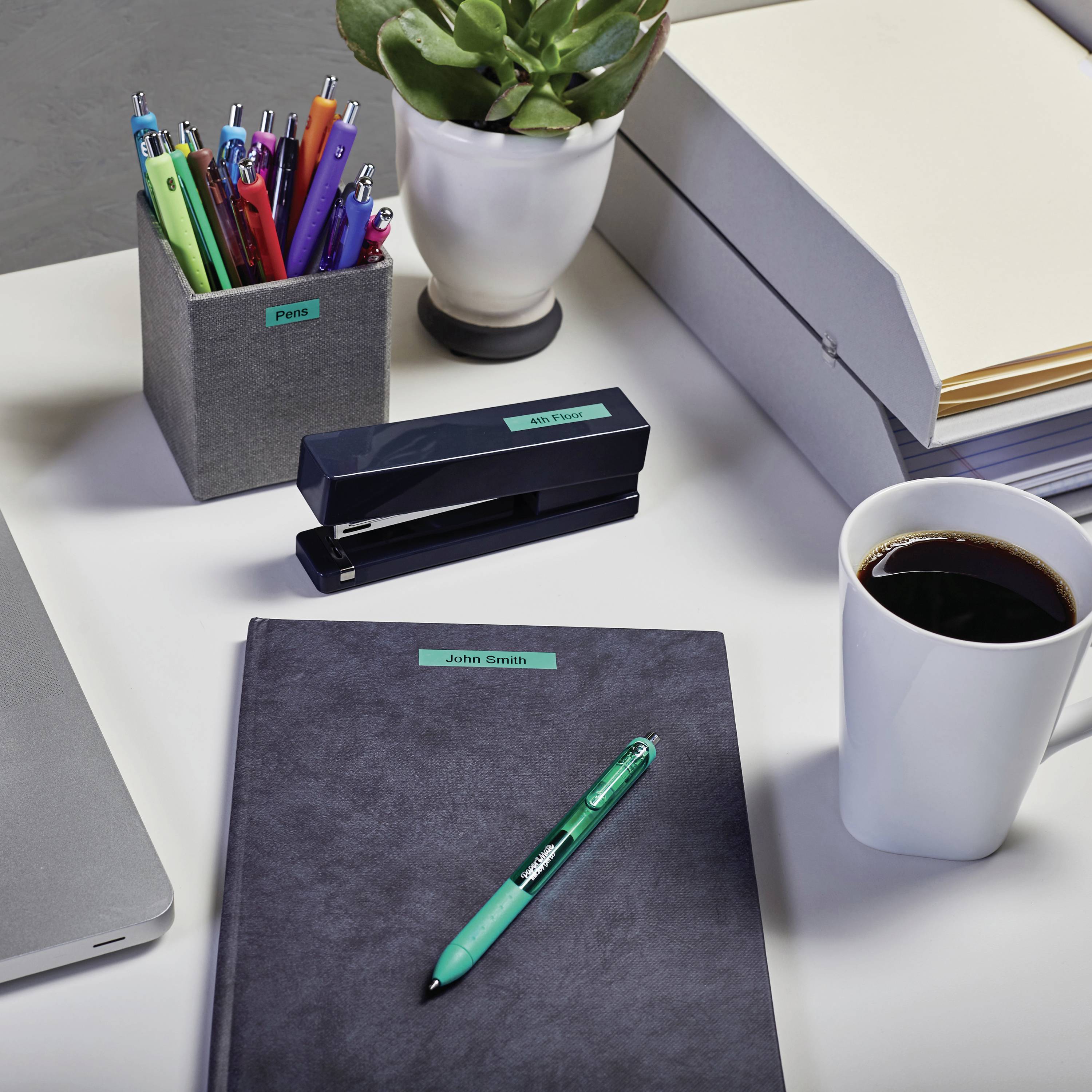 Ein Schreibtisch mit bunten Stiften, einer Pflanze, Ordnern, einer Kaffeetasse und einem Notizbuch mit einem grünen Stift darauf.