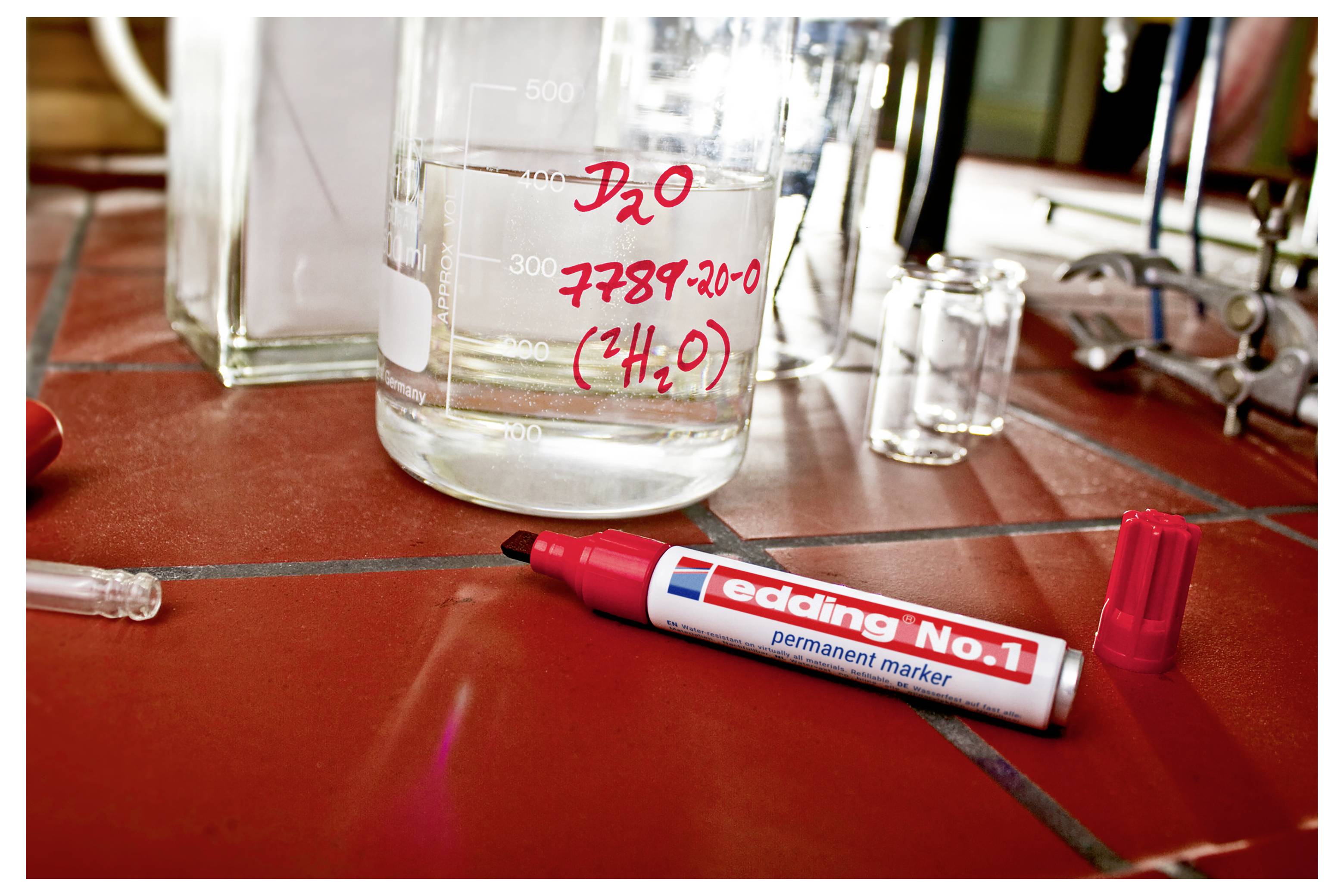 Ein roter permanenter Marker liegt auf einer gefliesten Labortischplatte neben einem Glasbecher mit der Aufschrift „D2O 7789-20-0 (H2O)".