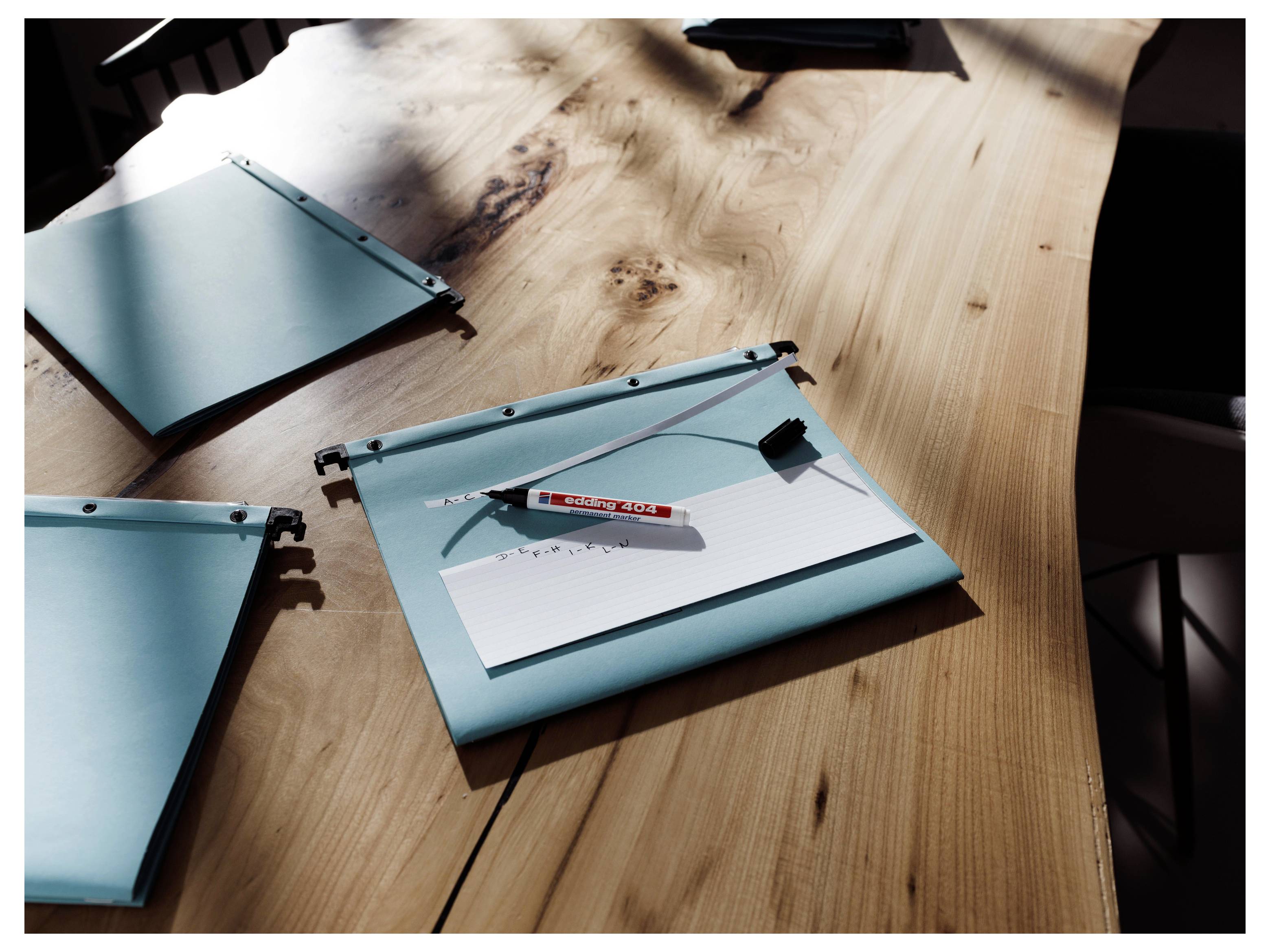 Drei blaue Ordner mit Papieren und ein Stift liegen verstreut auf einem Holzschreibtisch im hellen Sonnenlicht.