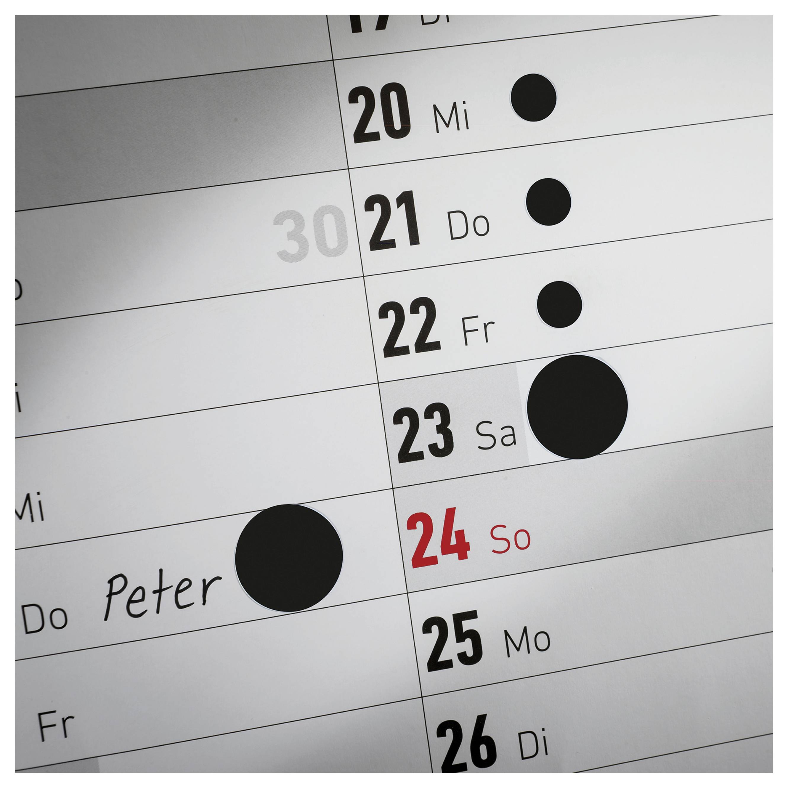 Nahaufnahme eines Wandkalenders mit den Tagen 20 bis 26; der 24. ist rot als Sonntag markiert, mehrere schwarze Punkte kennzeichnen Termine, einmal mit dem Namen 'Peter'.