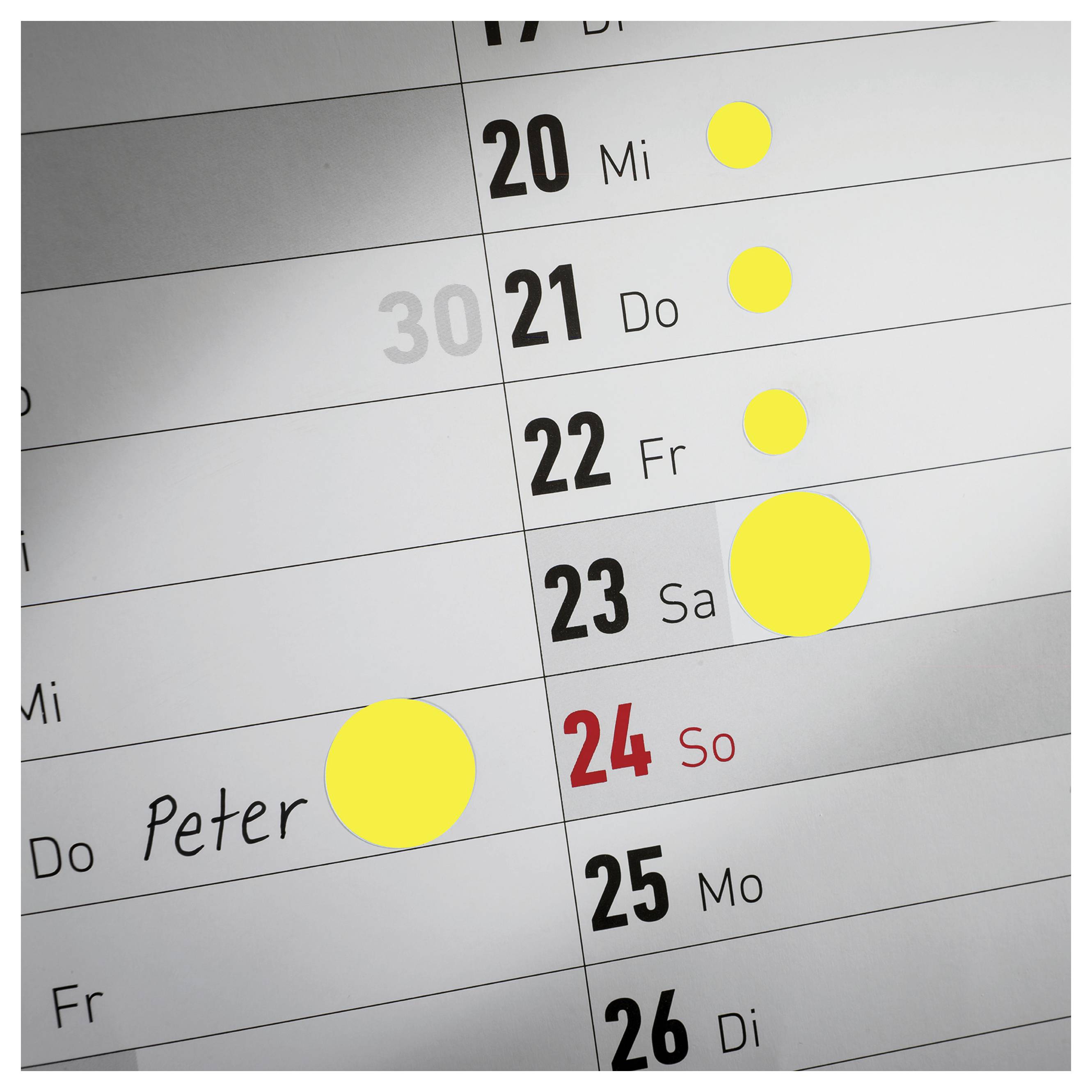 Nahaufnahme eines Wandkalenders mit den Daten 20 bis 26; mehrere gelbe Markierungen heben Termine hervor, ein Eintrag 'Peter' steht neben dem 24. Sonntag.