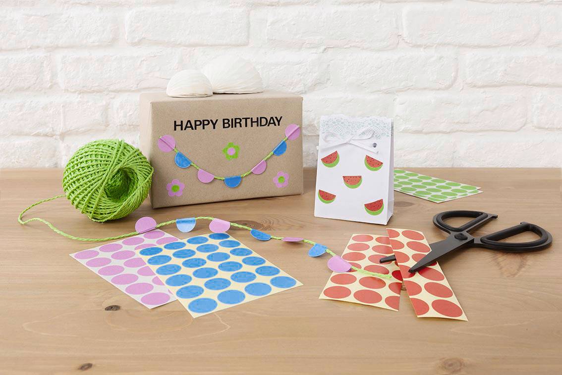Geschenkverpackung mit 'Happy Birthday', grüner Wolle, Schere und bunten Aufklebern auf einem Tisch vor einer weißen Ziegelwand.