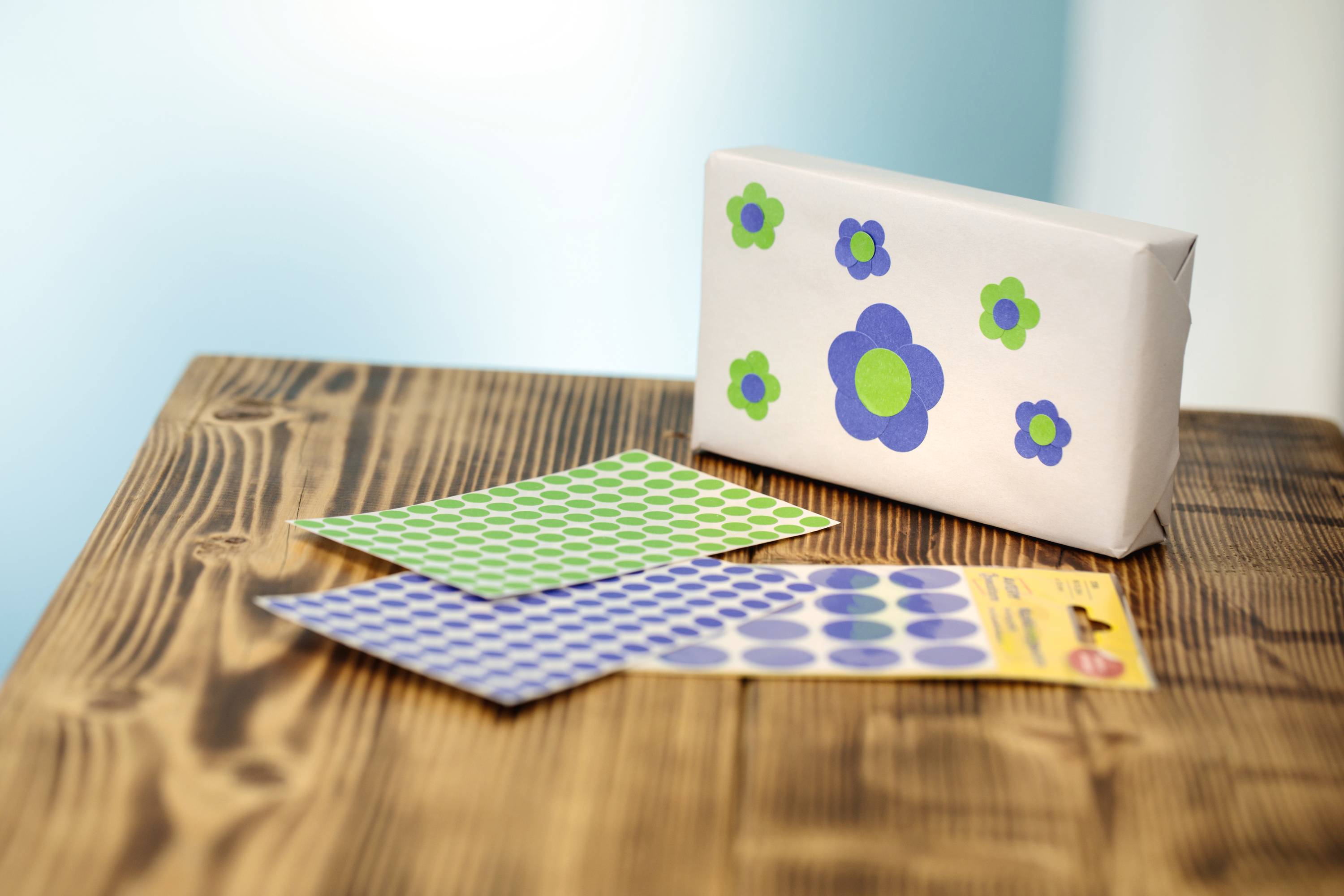 Geschenk mit Blumenmuster auf Holztisch, daneben grüne und blaue Klebepunktblätter.