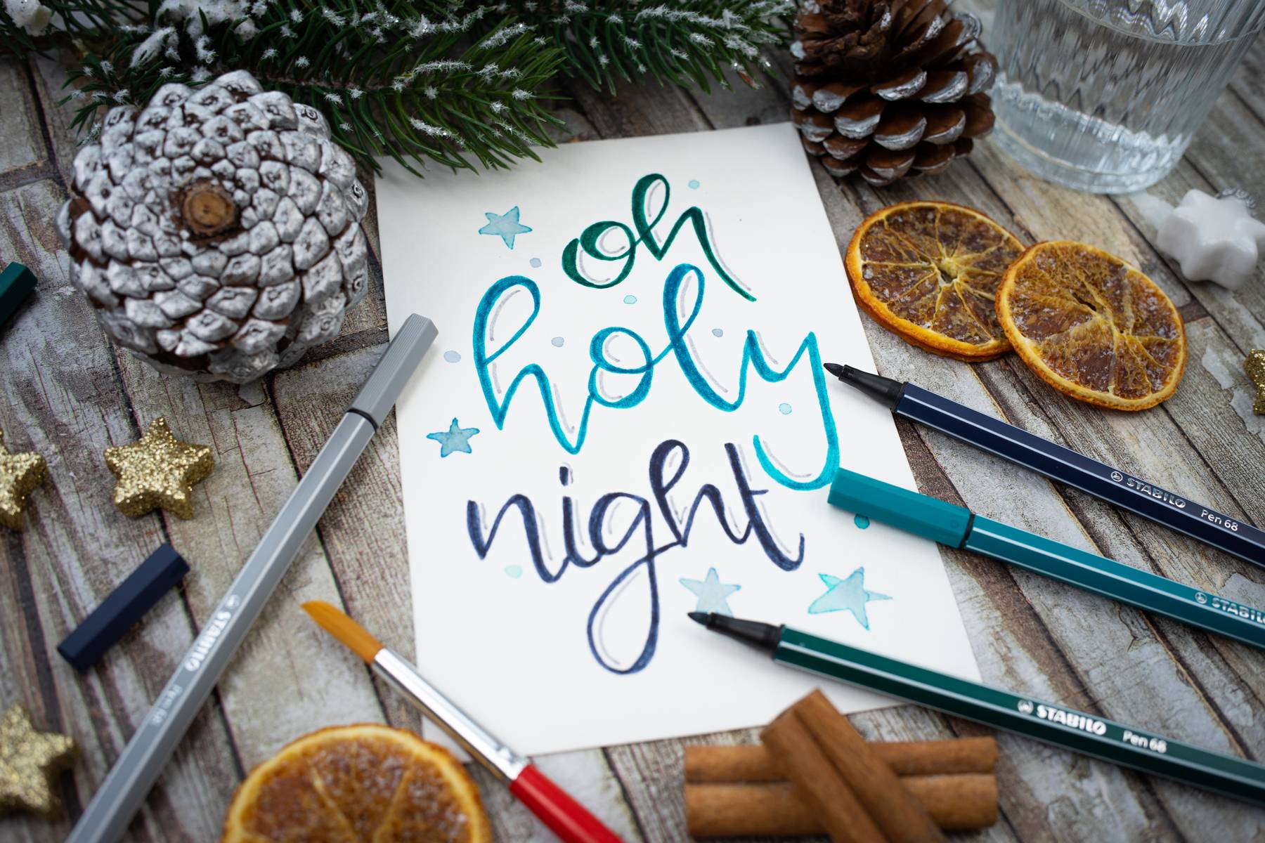 Handgeschriebene Karte mit 'oh holy night', umgeben von Tannenzapfen, getrockneten Orangenscheiben, Stiften und Pinsel auf Holztisch.