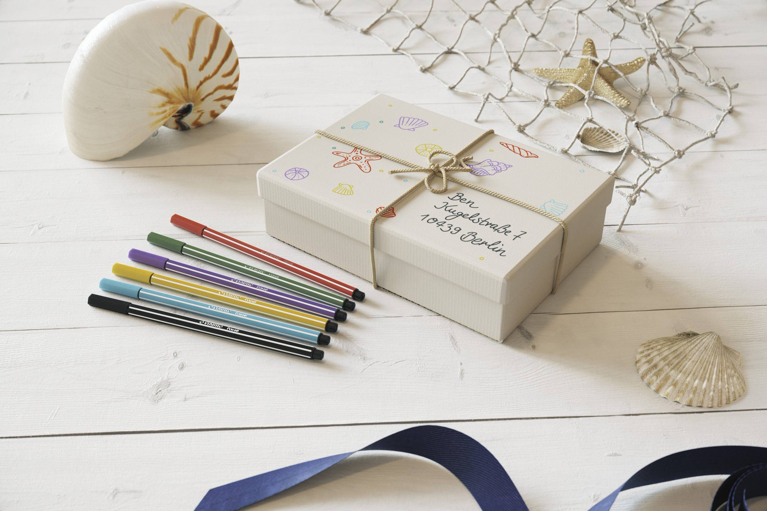 Eine dekorierte Geschenkbox mit der Aufschrift 'Zum Geburtstag!'. Daneben liegen bunte Stifte. Im Hintergrund sind Muscheln zu sehen.