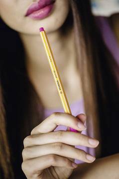 Eine Person hält einen gelben Bleistift mit leichtem Lächeln. Der Fokus liegt auf der Hand und dem Stift. Gesichtszüge sind unscharf.