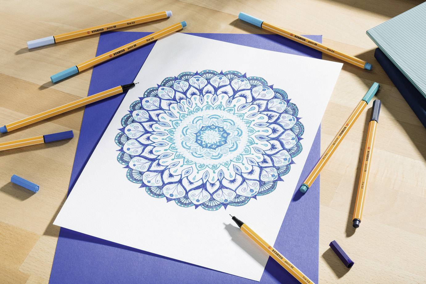 Mandala-Zeichnung in Blau- und Türkistönen auf einem Blatt Papier, umgeben von farbigen Stiften auf einem Holztisch.