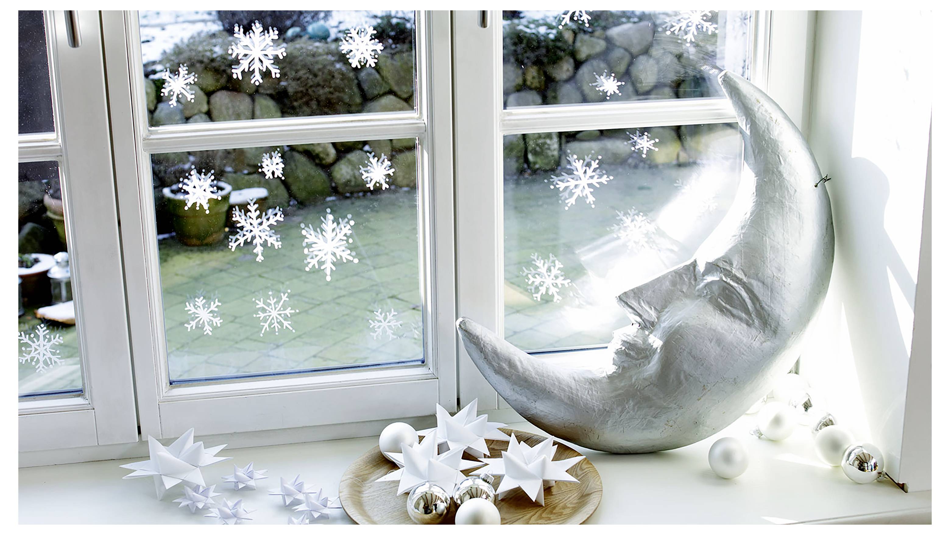 Weihnachtliche Schaufensterdekoration mit weißen Papierschneeflocken auf der Glasscheibe, einer silbernen Mondsichelskulptur und weißen Sterndekoration auf der Fensterbank.