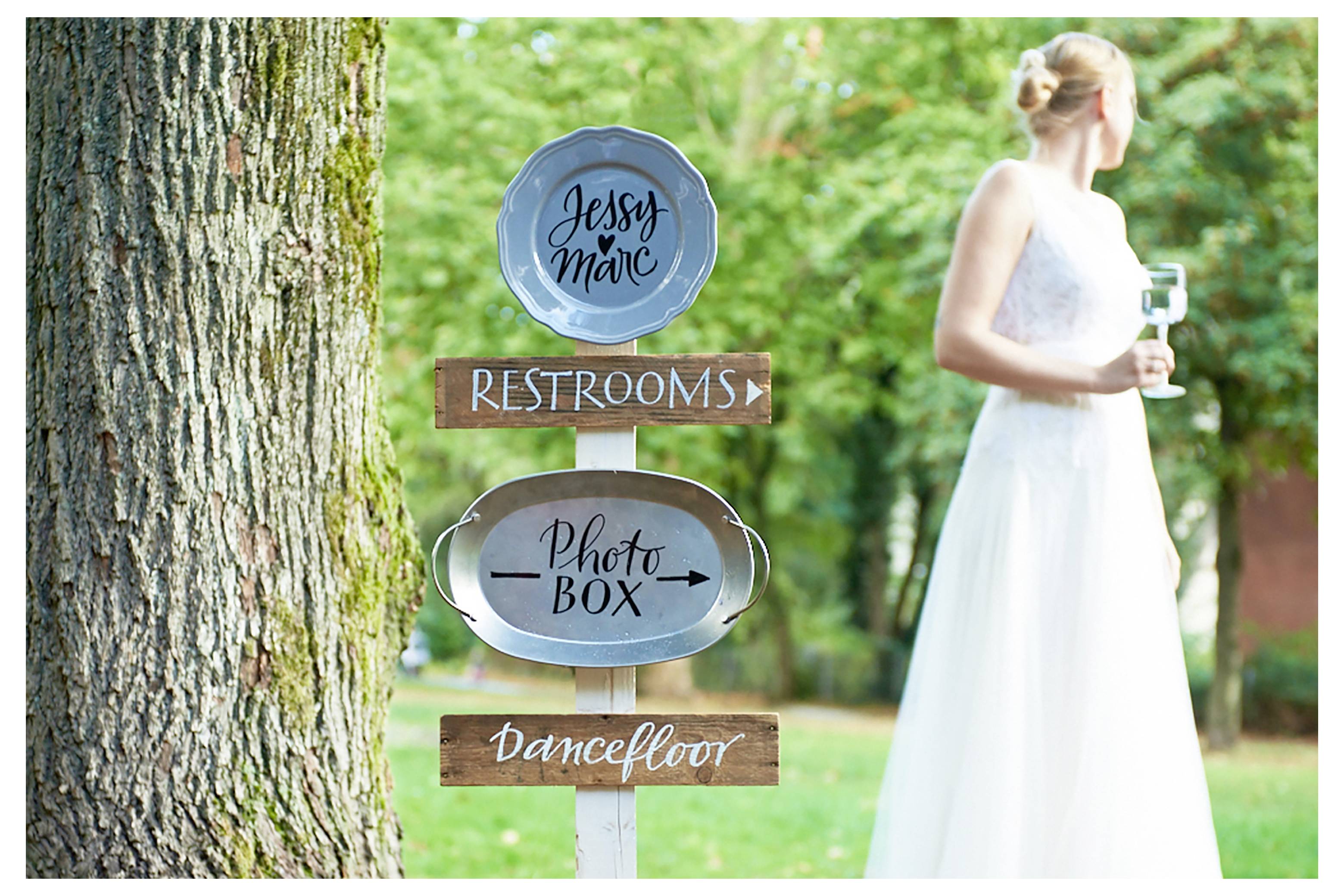 Hochzeitswegeschild mit Pfeilen für „TOILETTEN", „Fotokabine" und „Tanzfläche" in der Nähe einer Braut im weißen Kleid im Freien.