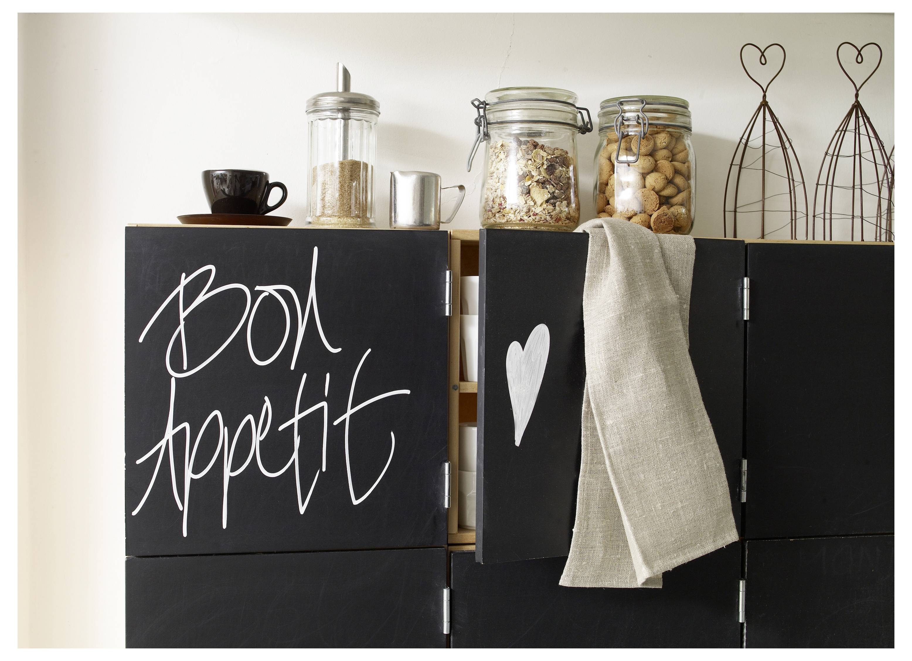 Schwarze Küchenschränke mit Tafelstil-Schriftzug „Bon appétit", ein weißes Herz, ein hängendes Handtuch und Glasgefäße oben.