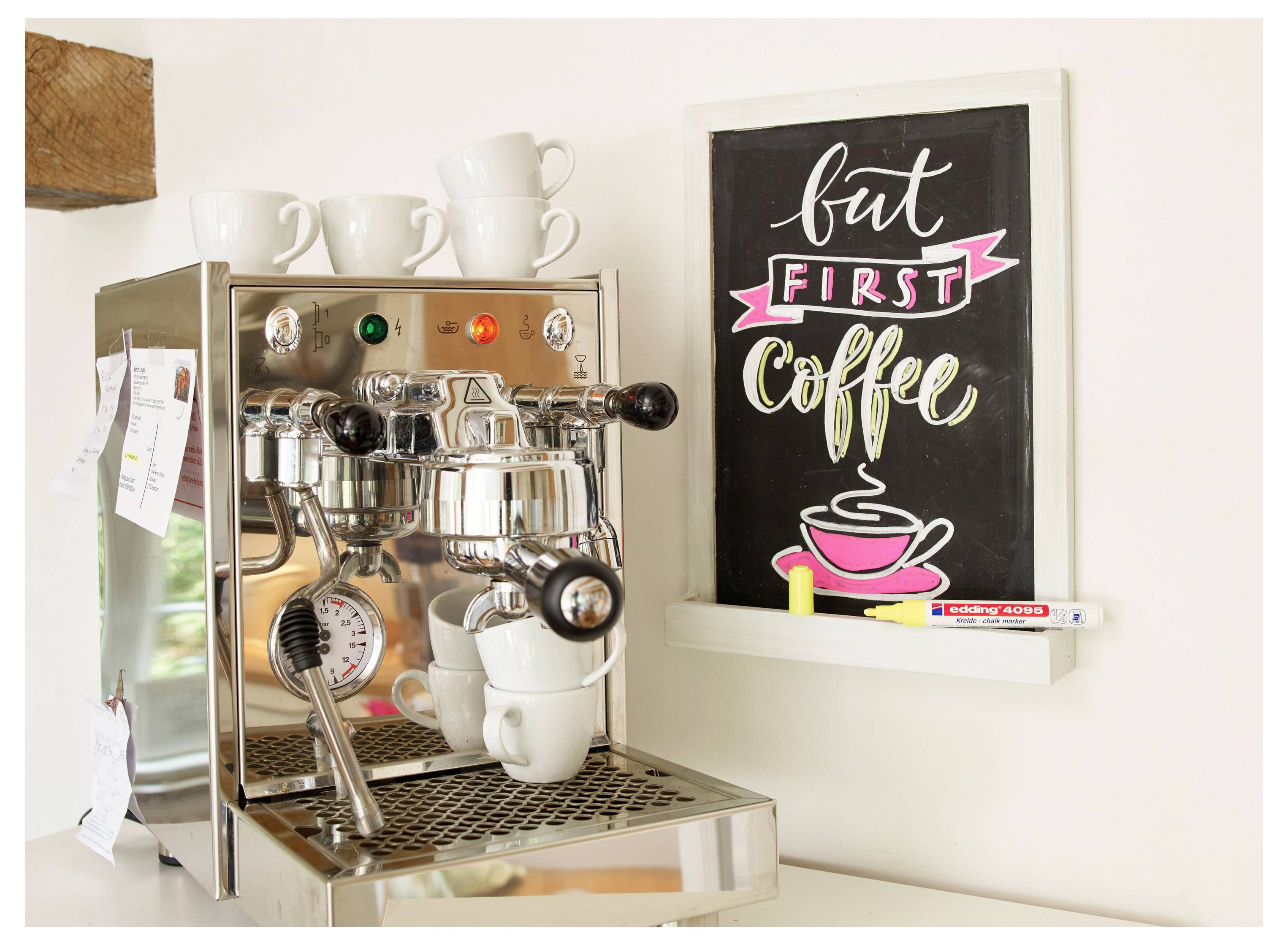 Espressomaschine mit Tassen und einem Wandschild mit der Aufschrift „aber ZUERST Kaffee" über einer rosa Kaffeetassenillustration.