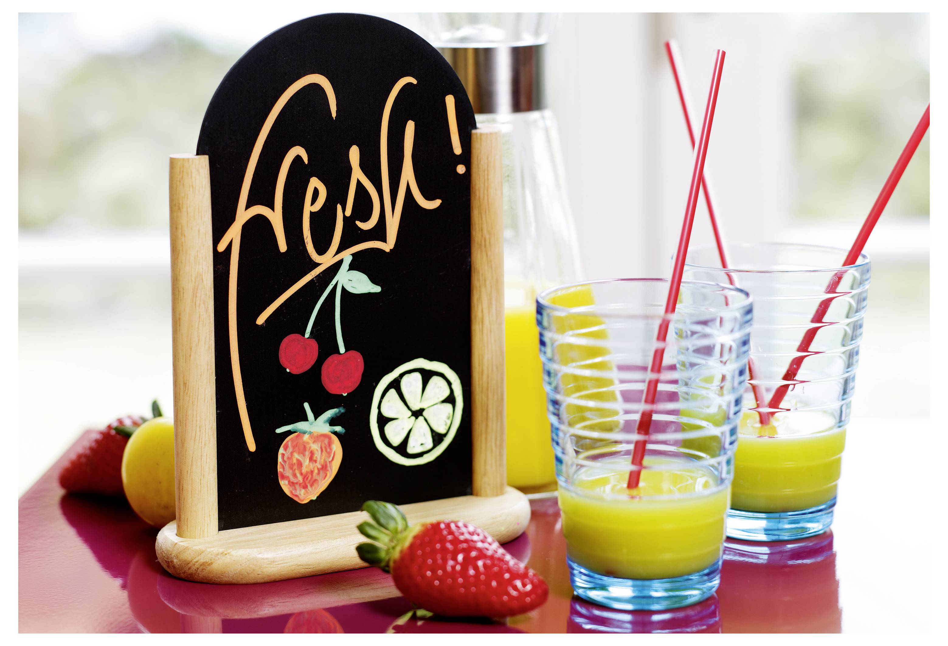 Frische Fruchtgetränkeanzeige mit Tafelschild mit der Aufschrift „Frisch", Zeichnungen von Kirschen, Erdbeere und Zitrusfrüchten, sowie zwei Gläser Saft mit roten Strohhalmen und Obst auf dem Tisch.
