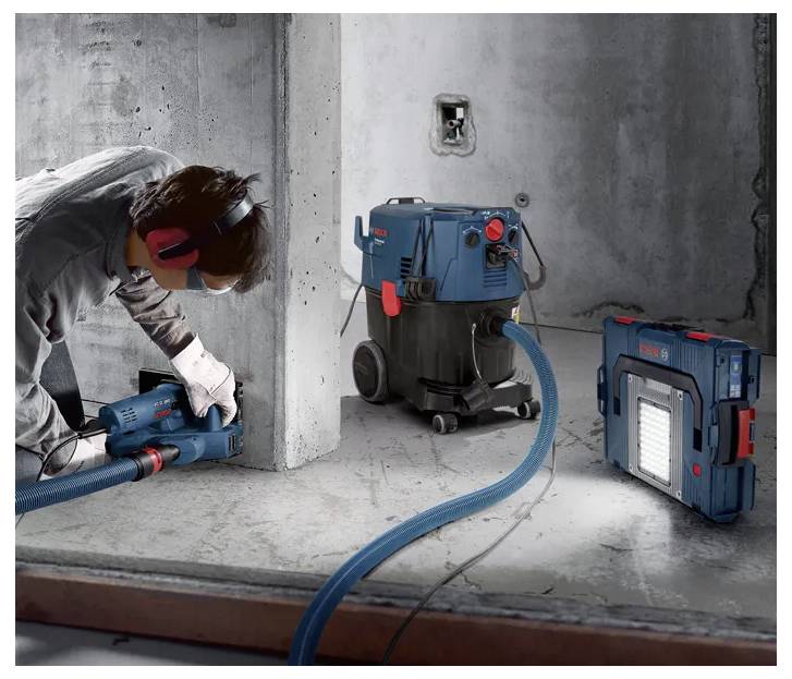 Arbeiter, der ein blaues Bosch-Elektrowerkzeug zum Schneiden von Beton in der Nähe einer Wand verwendet, mit einem Bosch-Staubabscheider und einer Arbeitsleuchte in einem unfertigen Raum.
