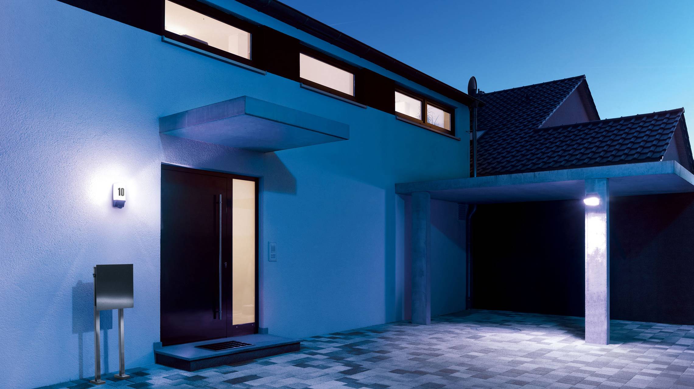 Einfamilienhaus bei Dämmerung, moderne Architektur mit flachem Dach, beleuchteter Eingang und Carport, blaue Beleuchtung umgeben.