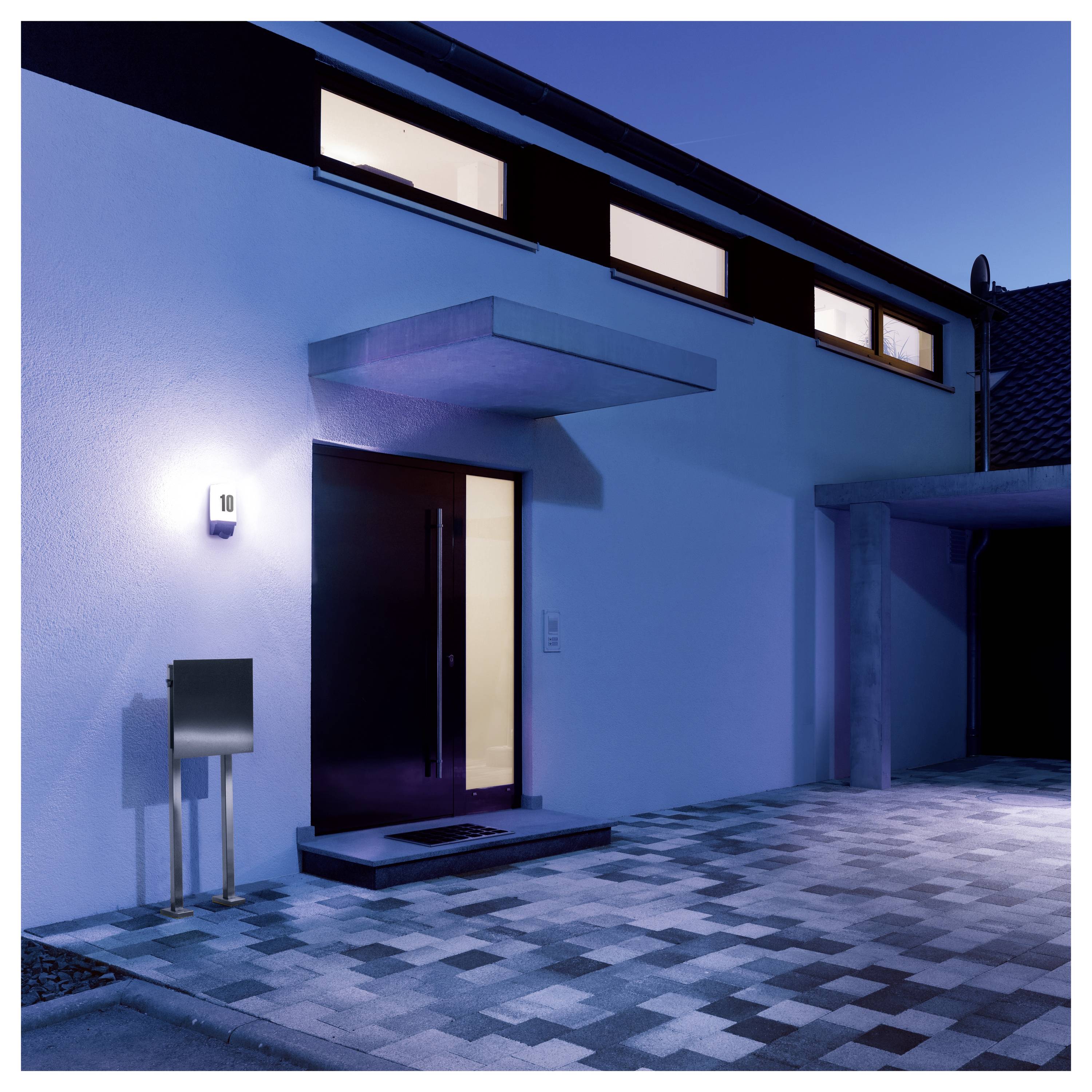 Moderner Hauseingang in der Abenddämmerung mit beleuchteter Haustür, Hausnummer 10 an der Wand und gepflasterter Einfahrt zur Garage
