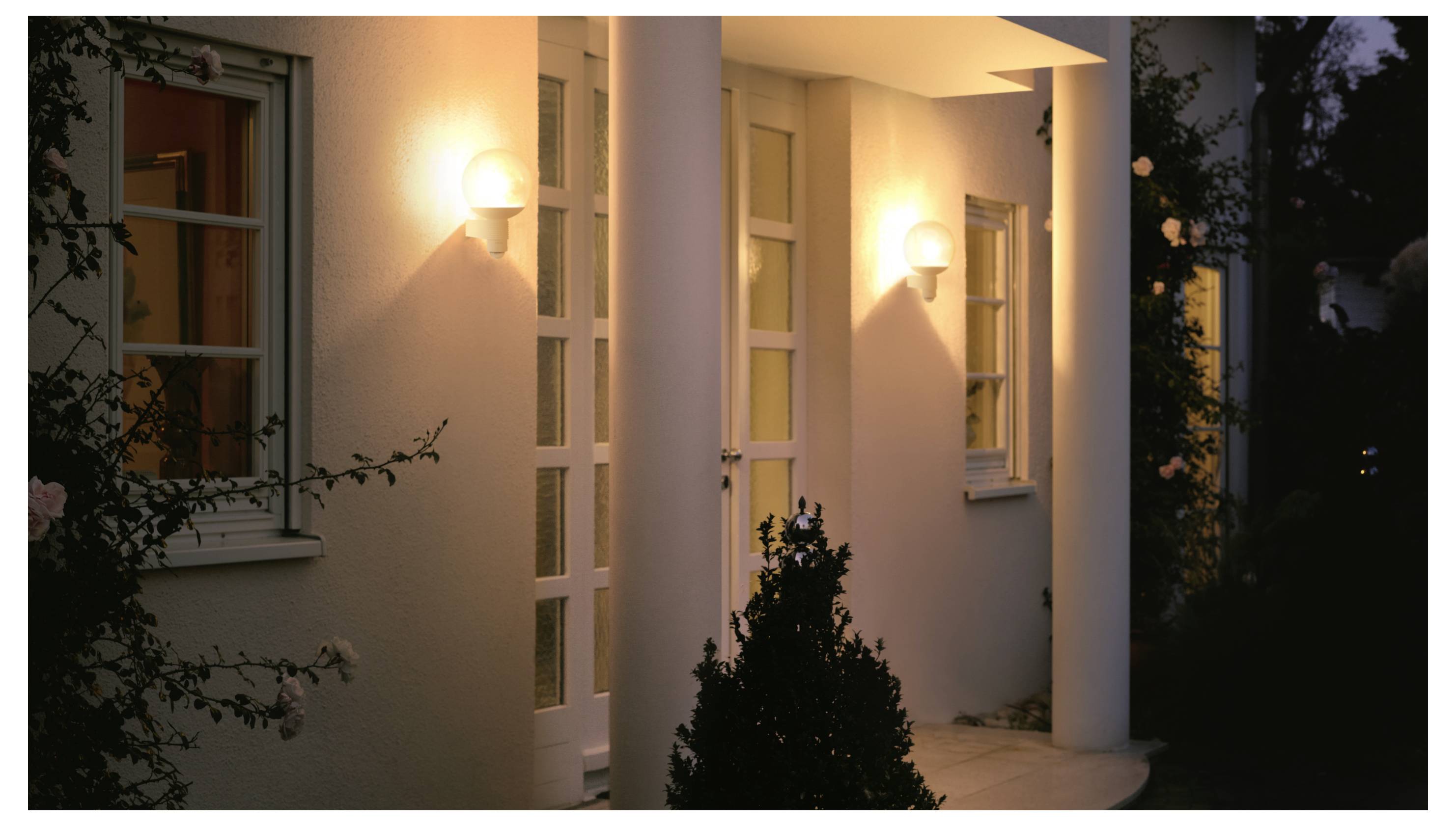 Abendliche Ansicht eines Hauseingangs mit leuchtenden Wandleuchten, weißen Säulen und einer Eingangstür, die von Fensterrahmen und Sträuchern umrahmt ist.