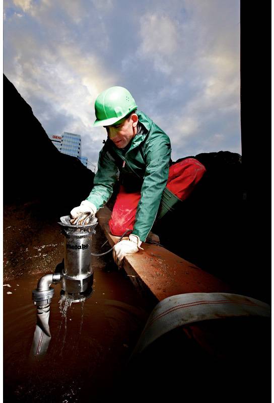 Eine Person mit Helm und Schutzkleidung arbeitet im Freien an einer Wasserpumpe in einer Baustelle. Wolken sind im Hintergrund sichtbar.
