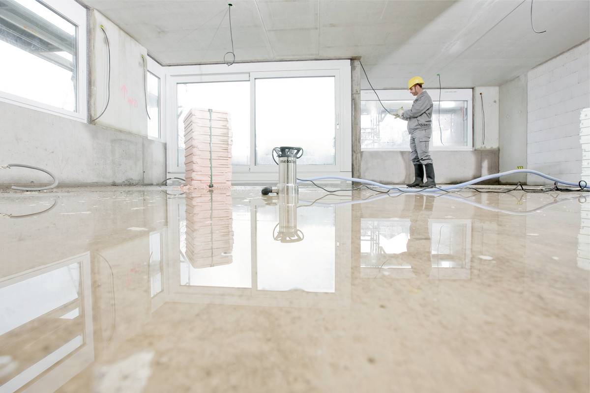 Ein Bauarbeiter in Schutzkleidung und Helm überprüft eine Baustelle mit großen Fenstern. Der Boden ist nass und reflektierend.