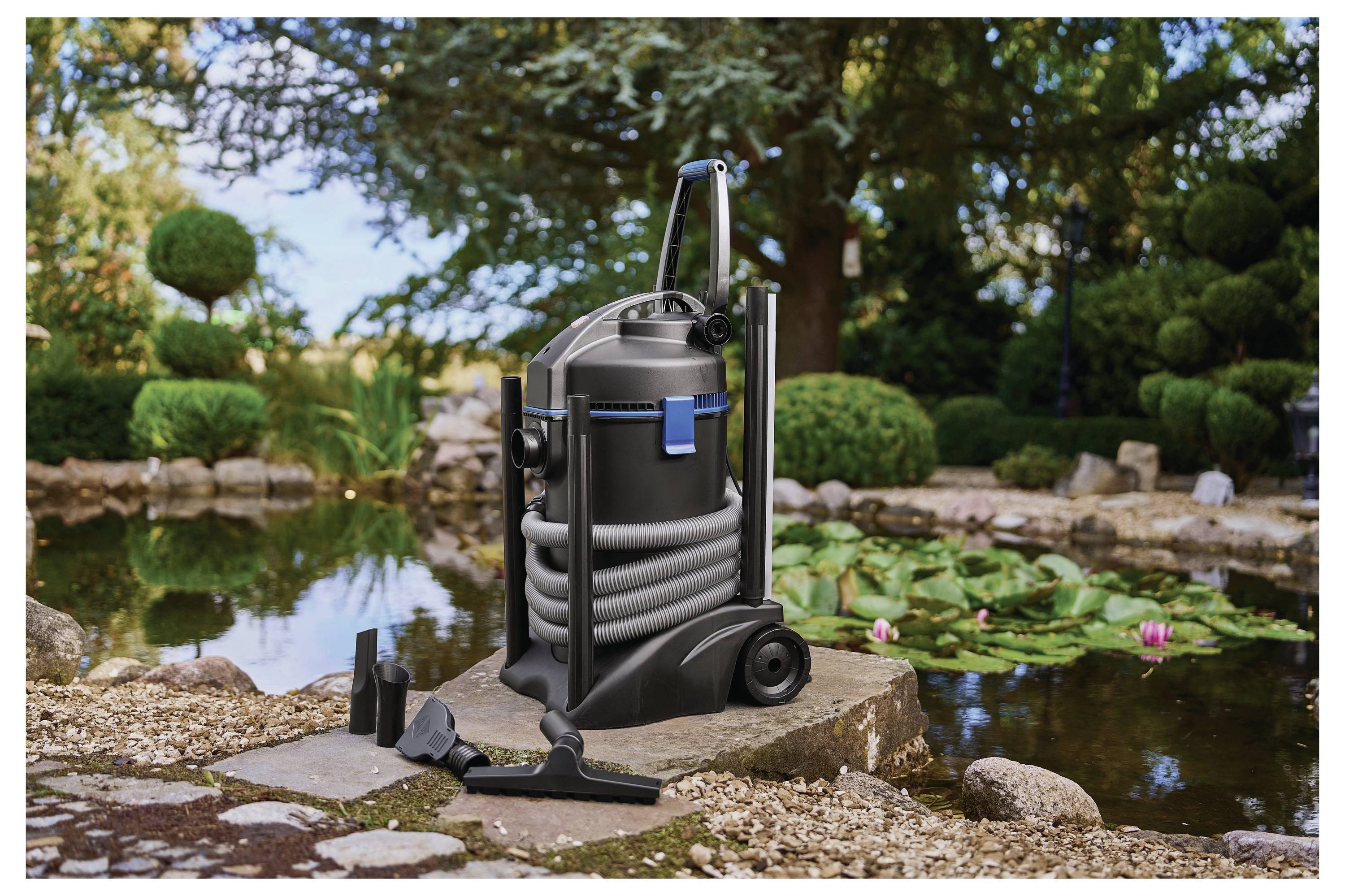Ein Teichstaubsauger auf einem Steinweg neben einem Gartenteich mit Seerosen, Steinen und üppiger Vegetation im Hintergrund.