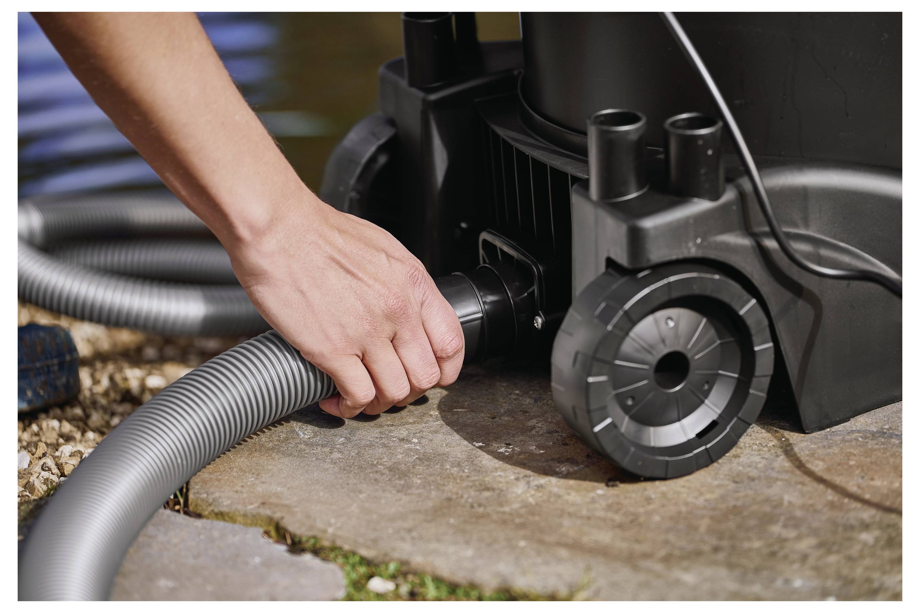 Eine Hand verbindet einen Schlauch mit einem Teichsauger auf einer Steinoberfläche und deutet damit auf die Reinigung oder Wartung eines Teichs hin.