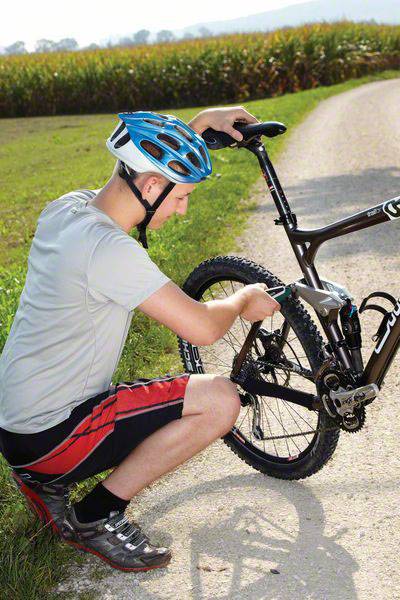 Eine Person trägt Helm und kniet auf einem Feldweg, während sie das Hinterrad eines Fahrrads repariert. Im Hintergrund ist ein Feld zu sehen.