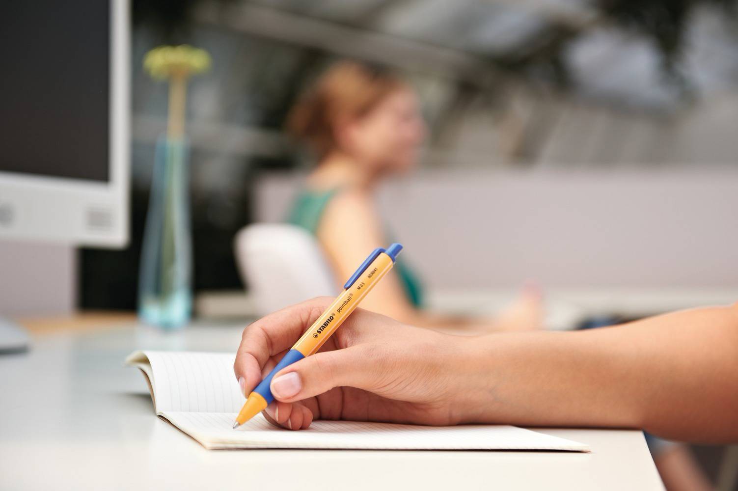 Eine Hand schreibt mit einem blauen und gelben Kugelschreiber in ein Notizbuch. Im Hintergrund ist unscharf eine Person zu sehen.